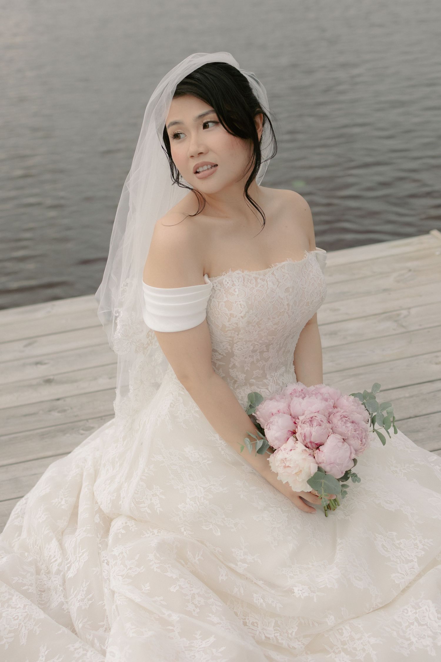 Asian bride in off-shoulder lace gown holding pink peony bouquet seated on a dock.