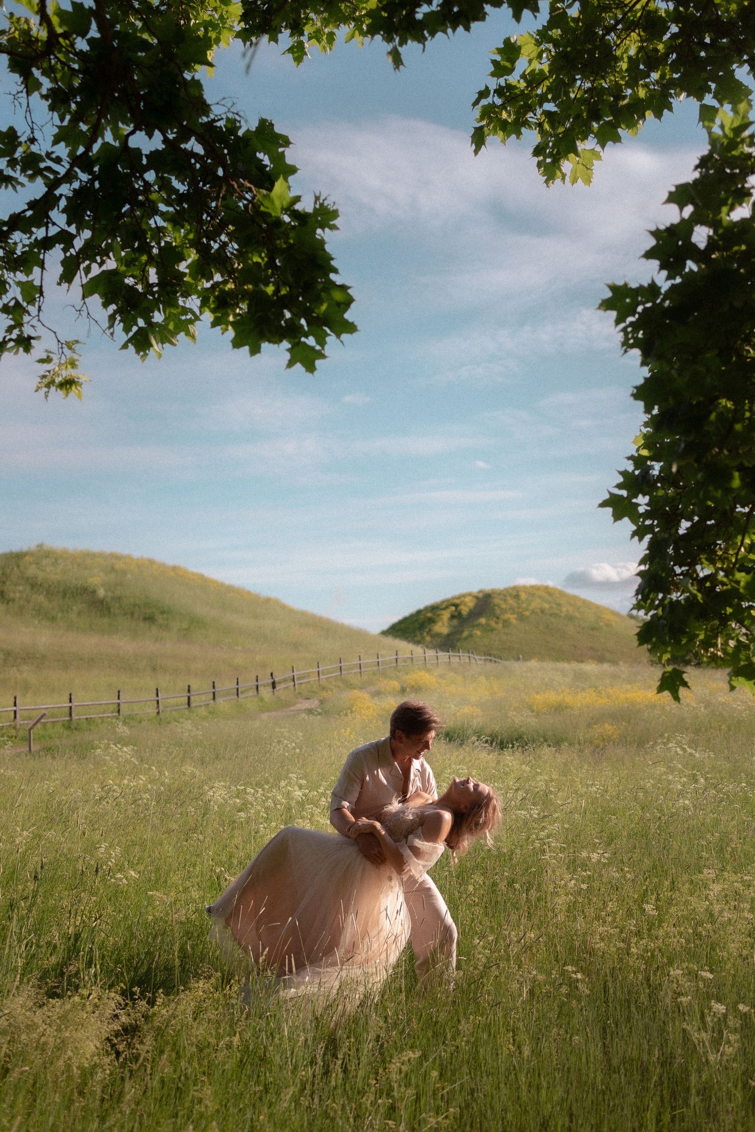 Couple dancing romantically in a lush green meadow with rolling hills and blue sky backdrop.