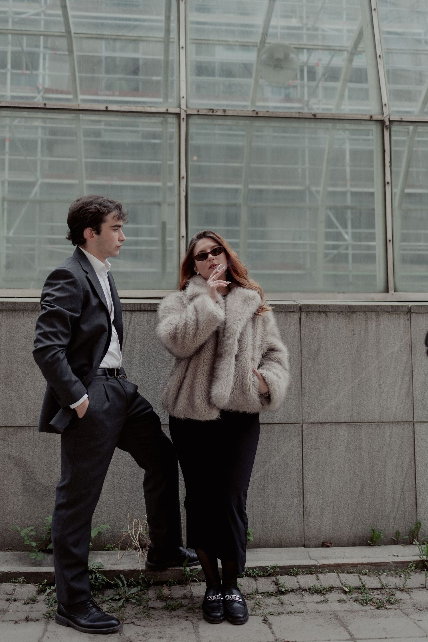 Stylish couple posing in front of glass greenhouse wall, woman in fur coat and sunglasses with suited man.