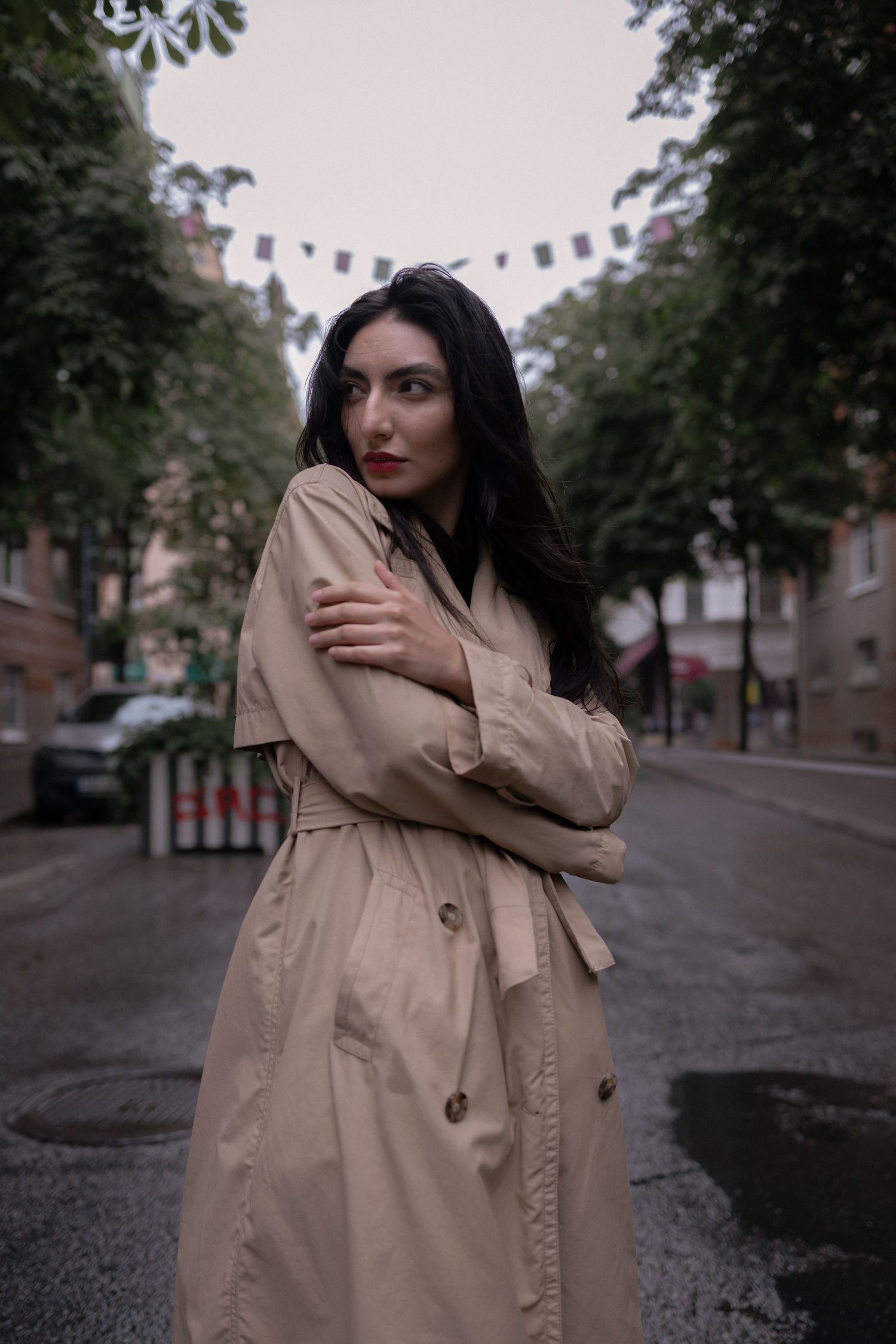 Dark-haired woman in beige trench coat on rainy urban street.