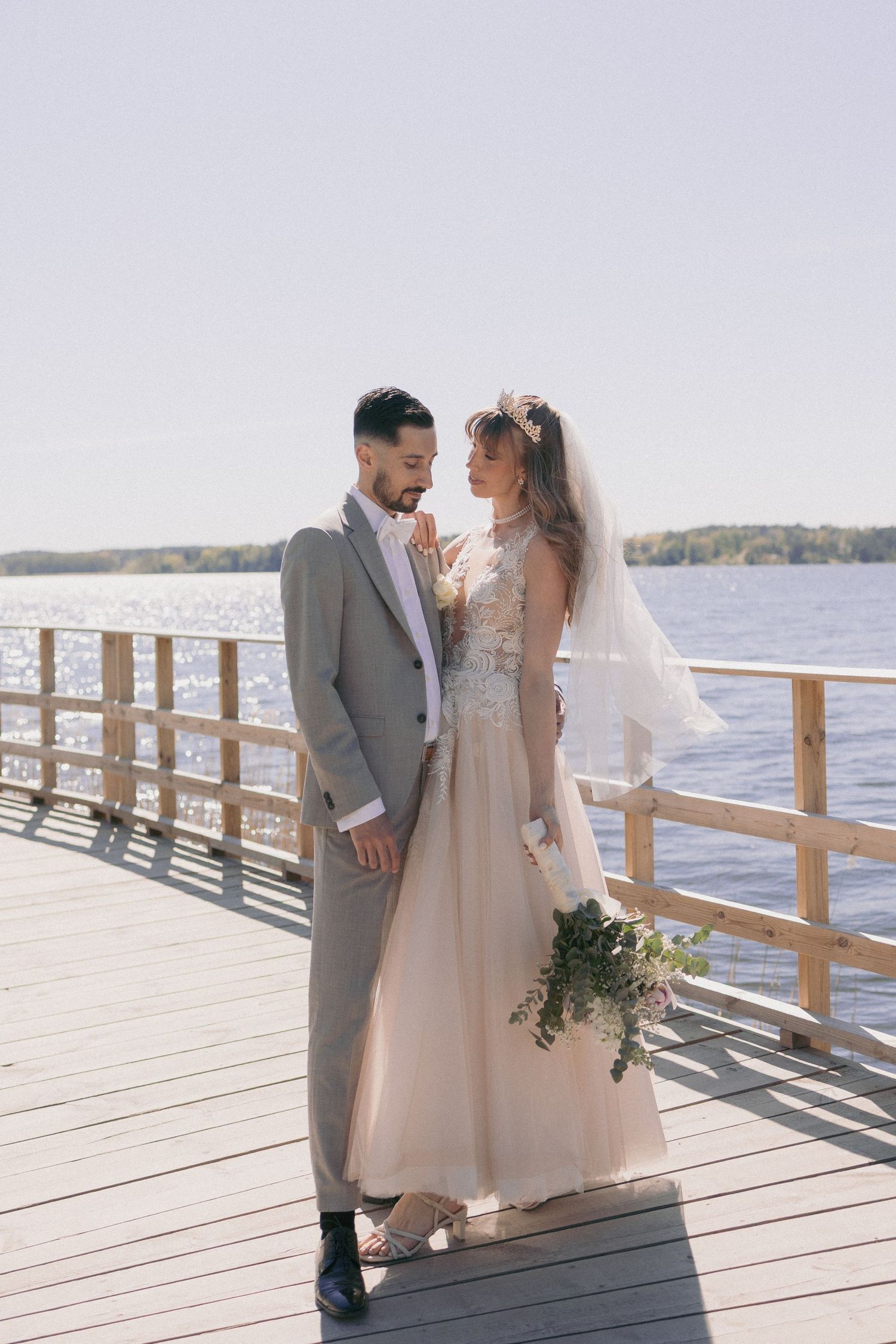 Bride and groom standing close on a sunny lakeside dock in elegant wedding attire.