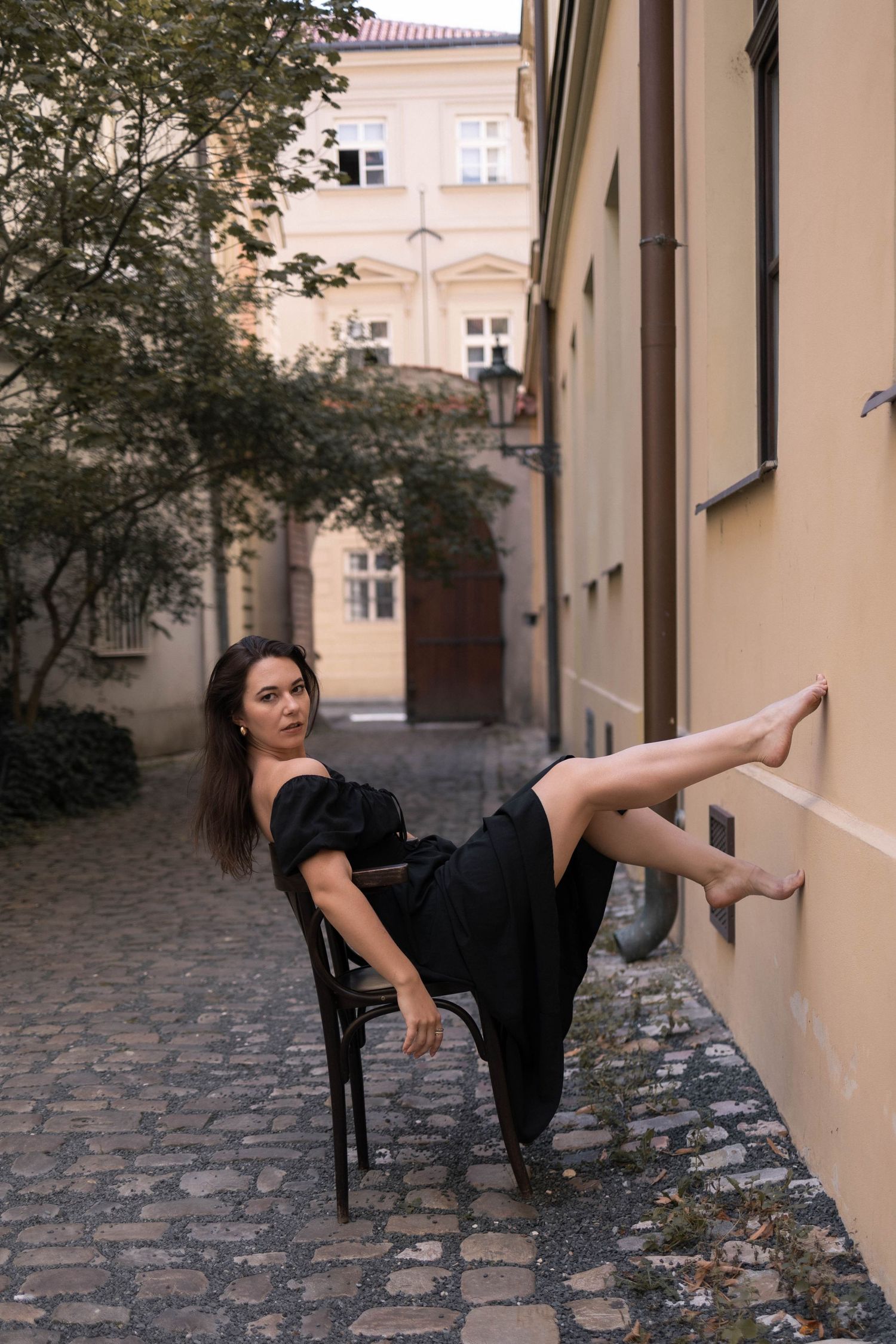Woman in black dress sits on chair with leg raised against wall in cobblestone alley.