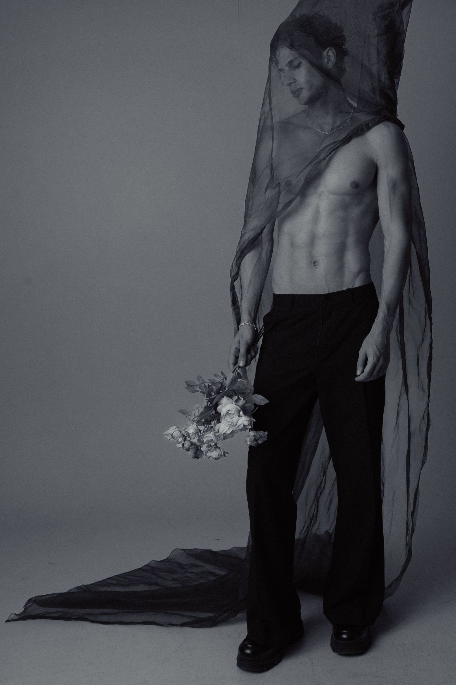 Shirtless man holding flowers draped in sheer veil in black and white photo.