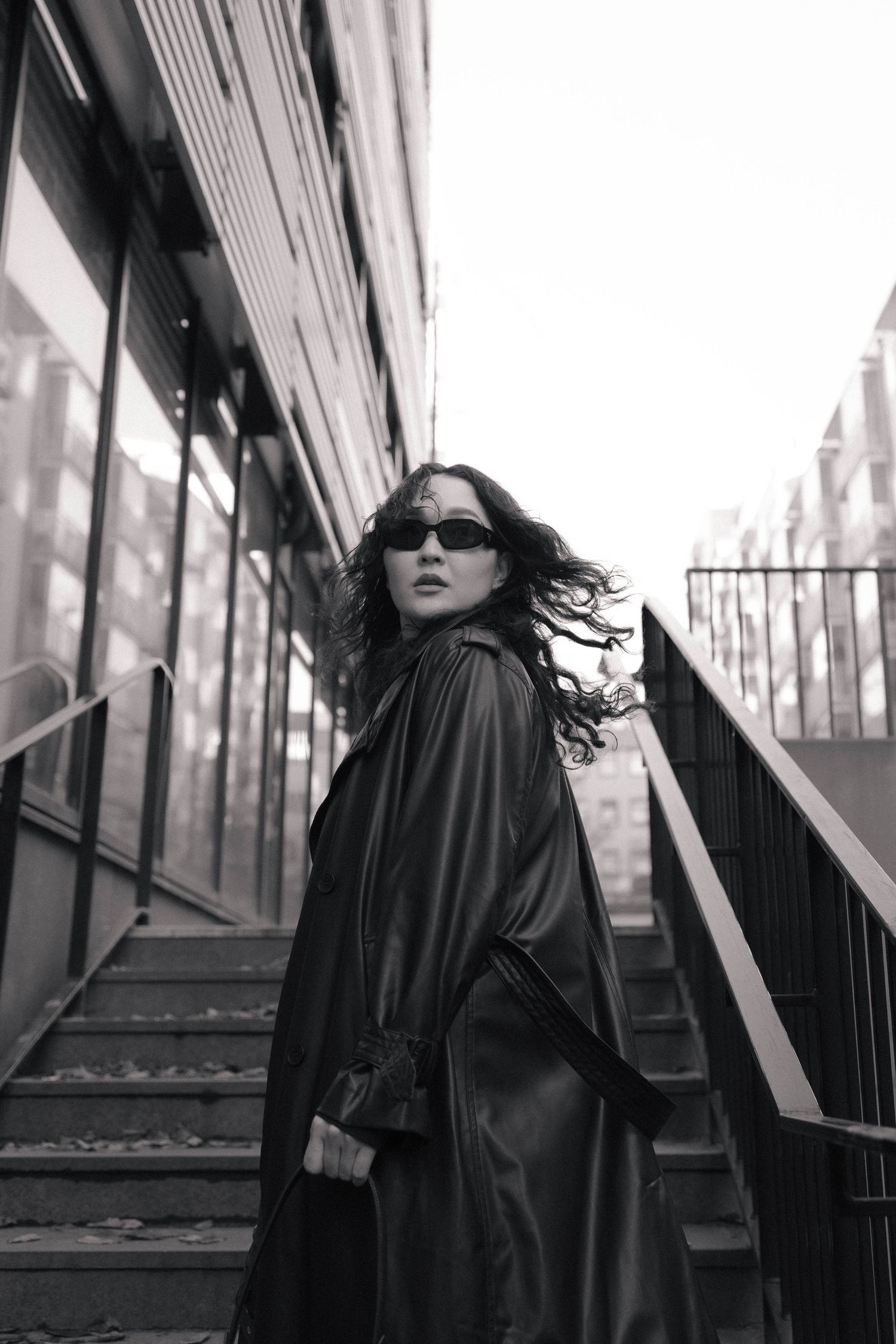 Black and white portrait of a woman in leather coat and sunglasses on urban staircase.