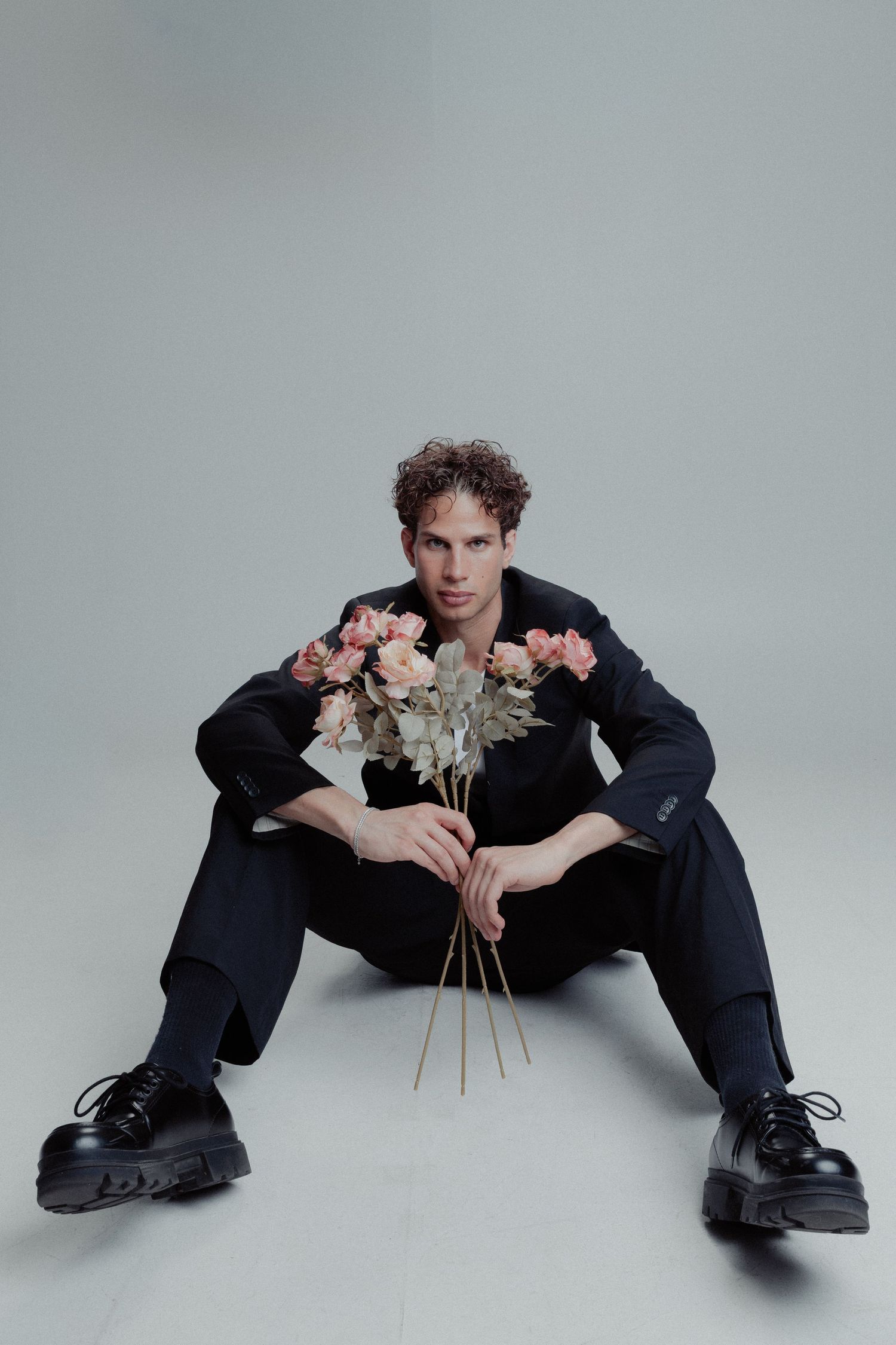 Man in black suit sitting on floor holding pink roses against a neutral grey background.