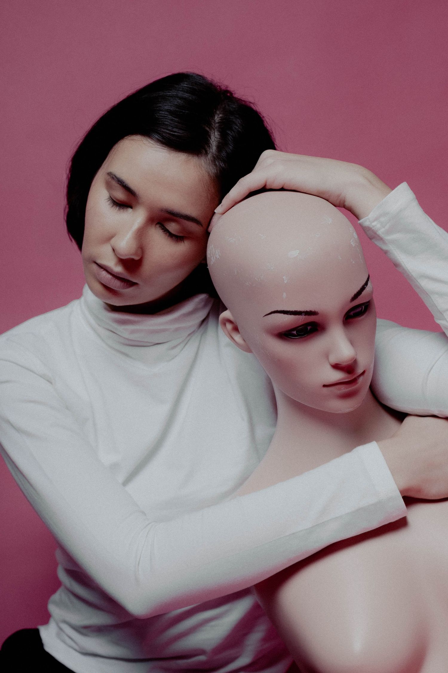 A dark-haired woman in a white turtleneck poses with a bald mannequin on a pink background.