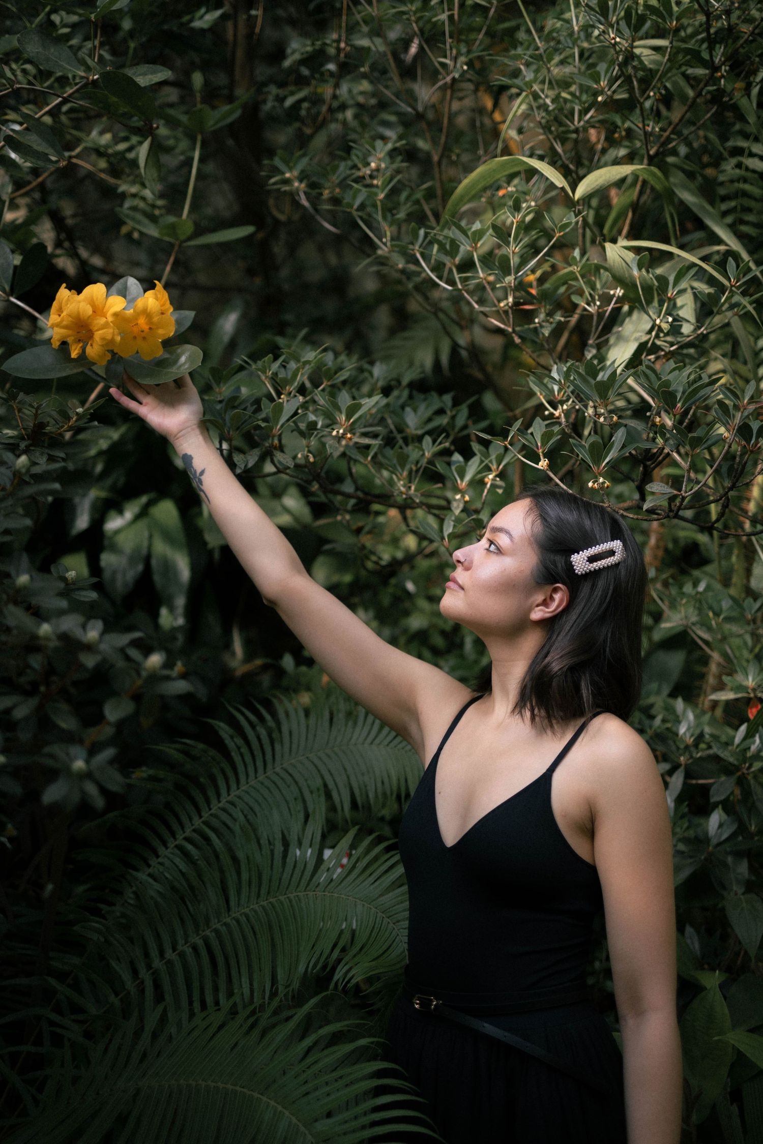 Woman in black top reaches up to touch yellow flowers among lush green tropical foliage.