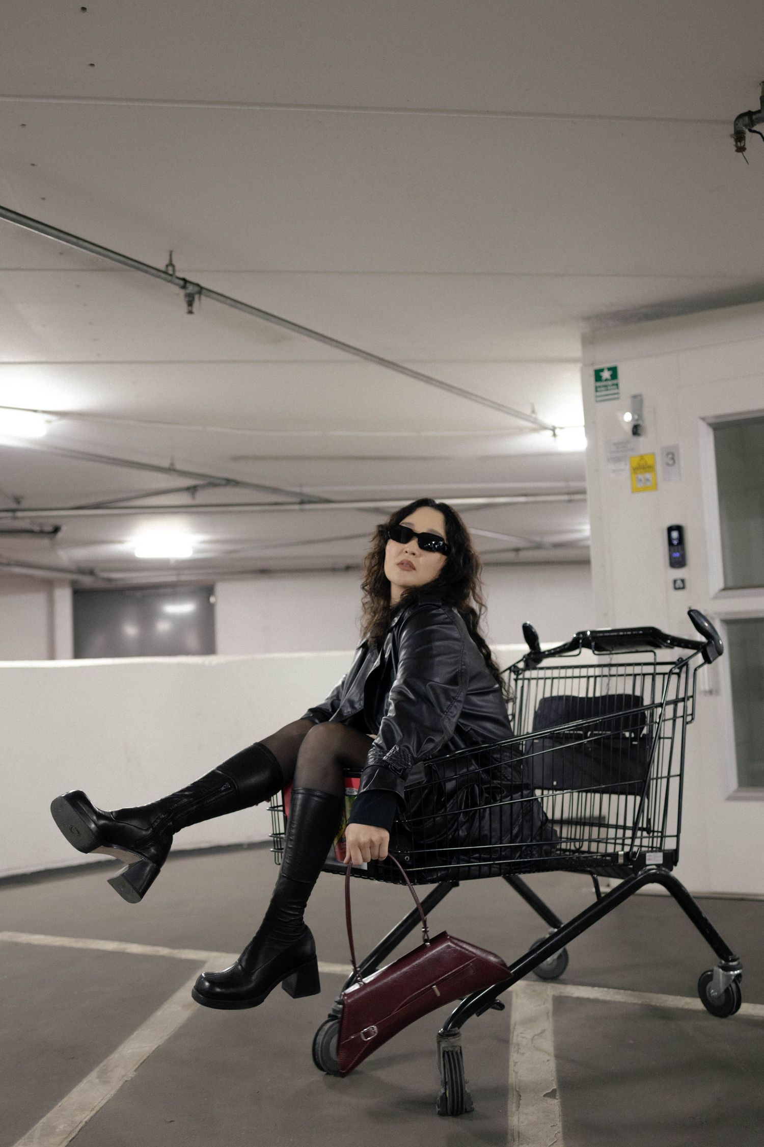 Woman in black leather jacket sitting in shopping cart in parking garage.