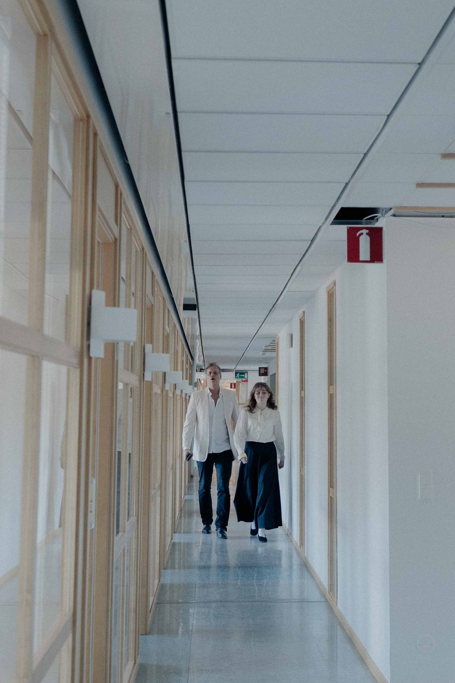 Two professionals walking down a bright modern office corridor with wooden doors and white walls.