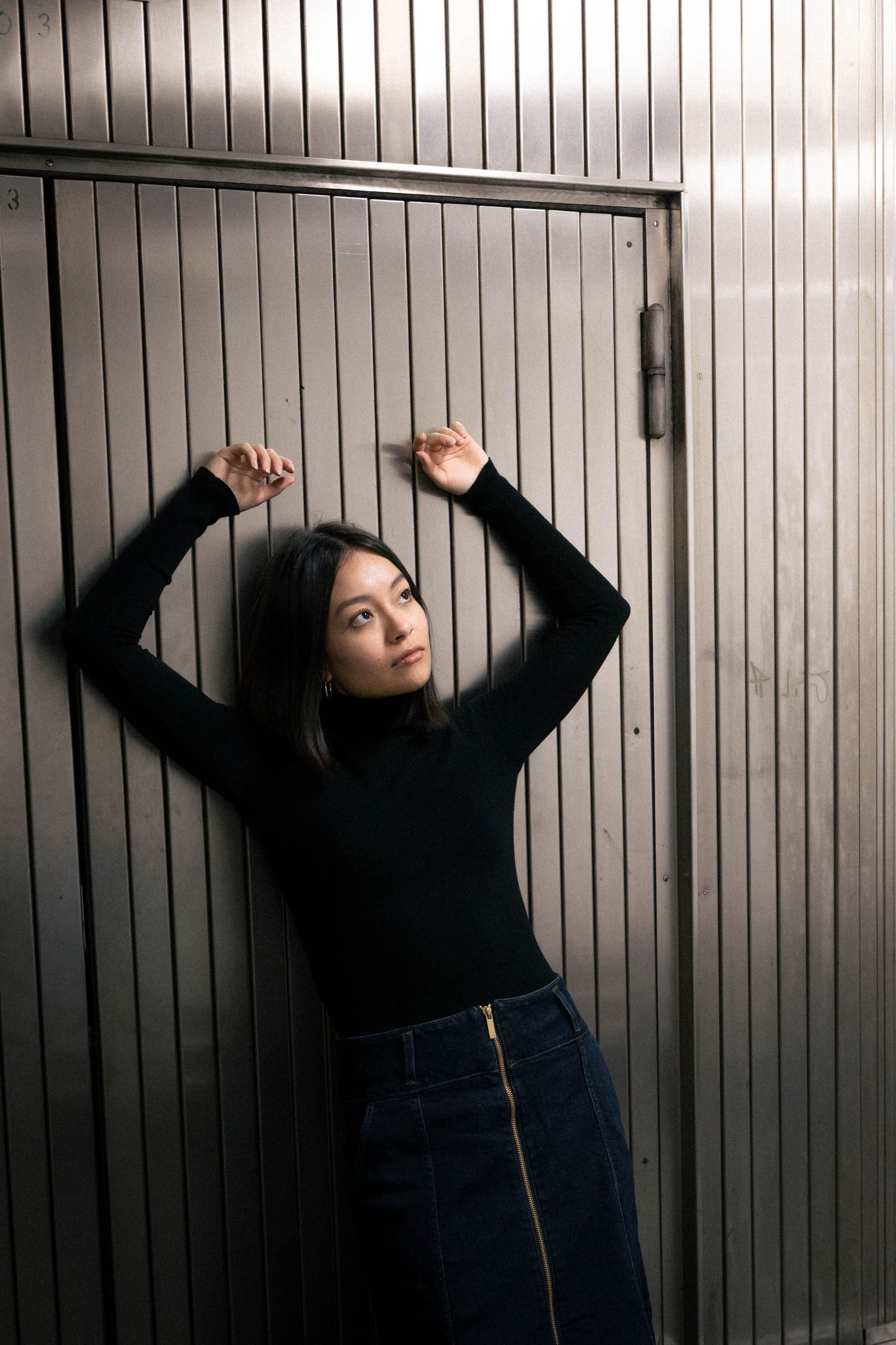 A young woman in a black turtleneck and denim skirt poses with arms raised against a metal door.