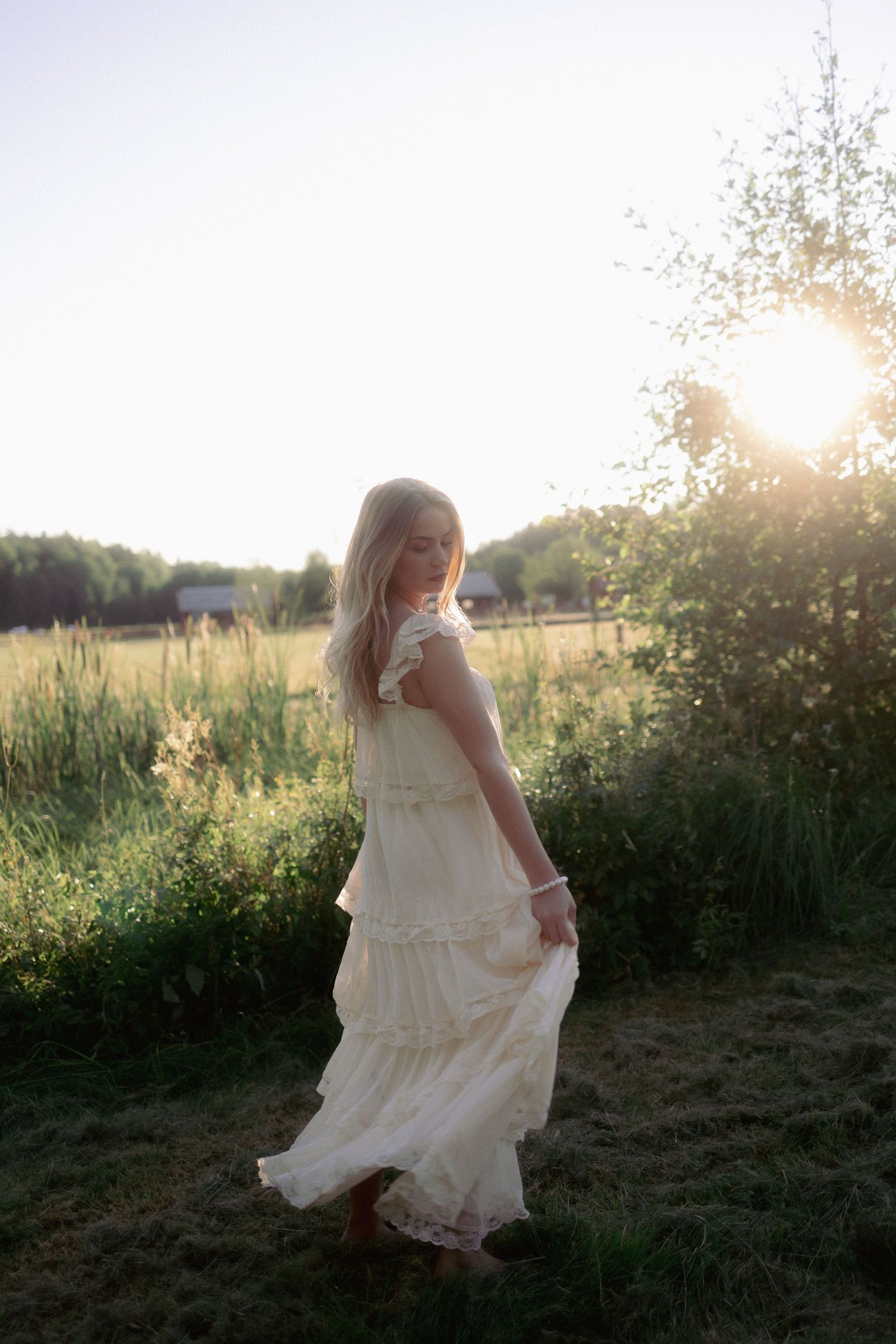 Blonde woman in white ruffled dress twirling in golden sunset light in a green field.