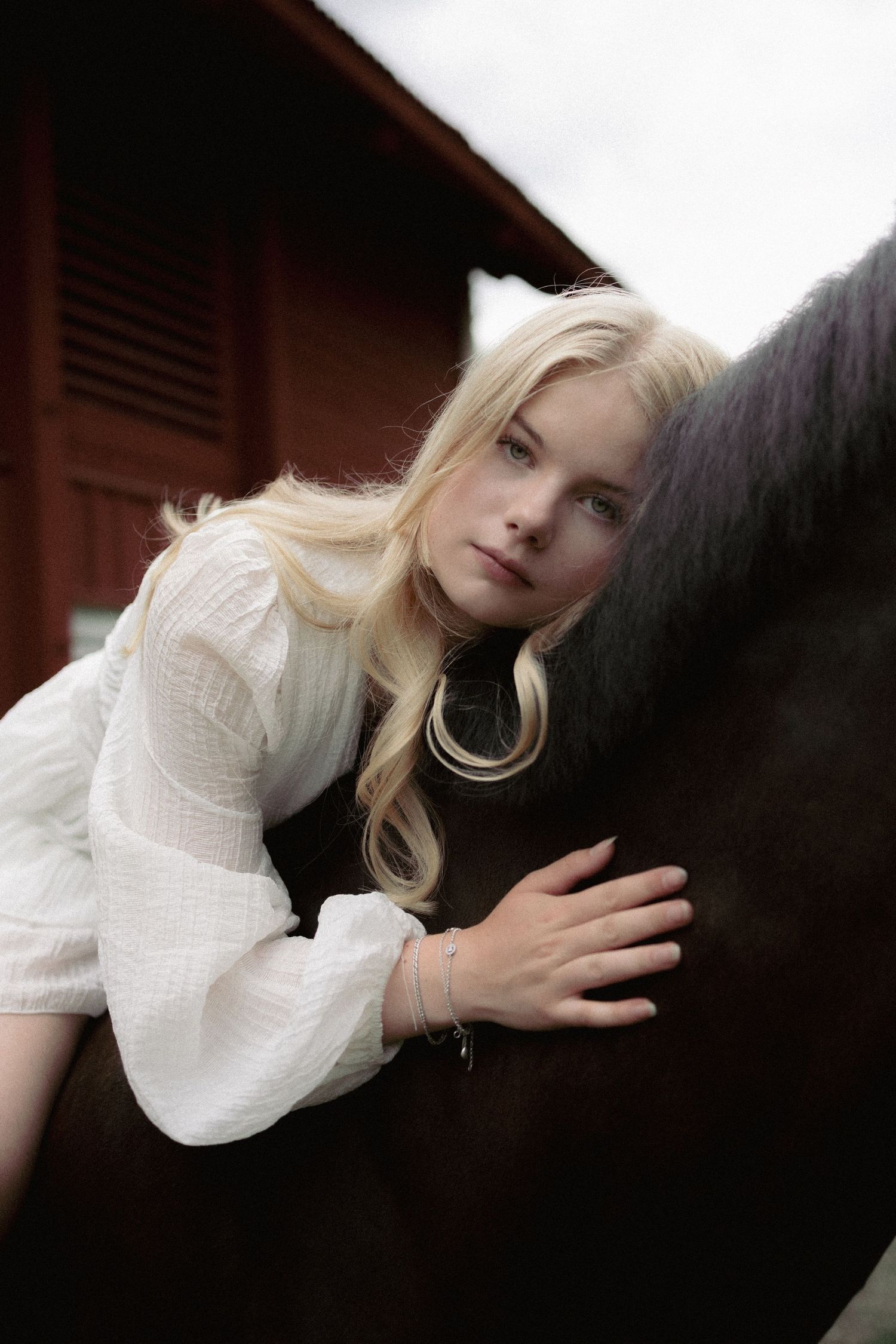 Blonde girl in white blouse tenderly embracing a dark horse near a red barn.