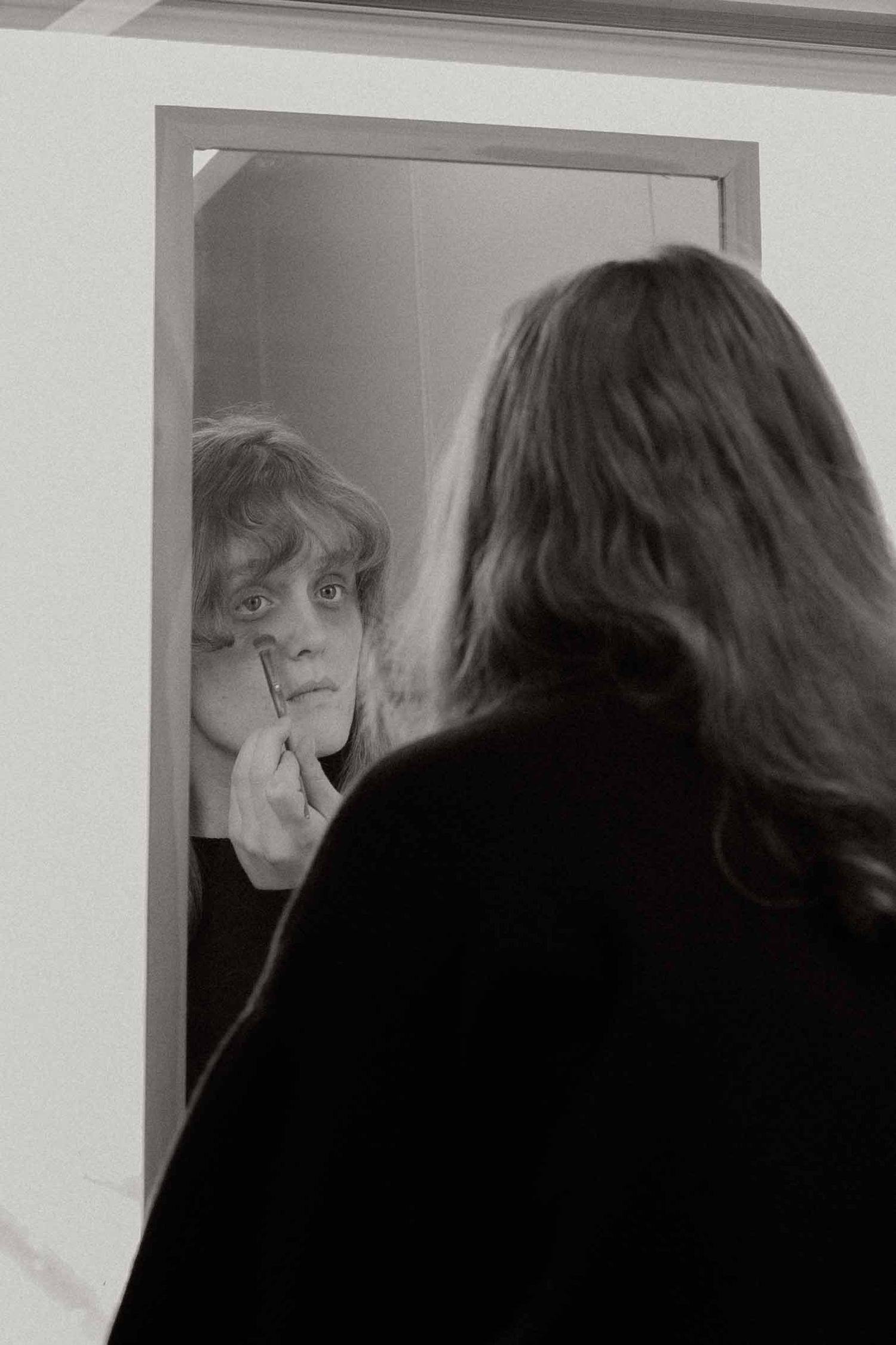Black and white photo of person applying makeup while looking into a mirror reflection.