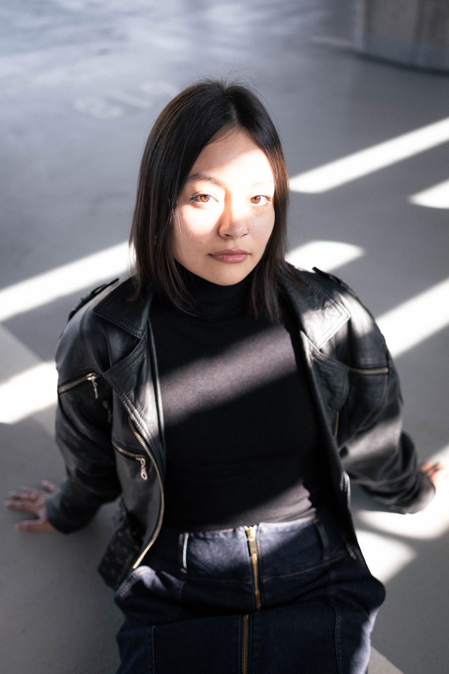 Asian woman in black leather jacket posing in sunlit industrial space.
