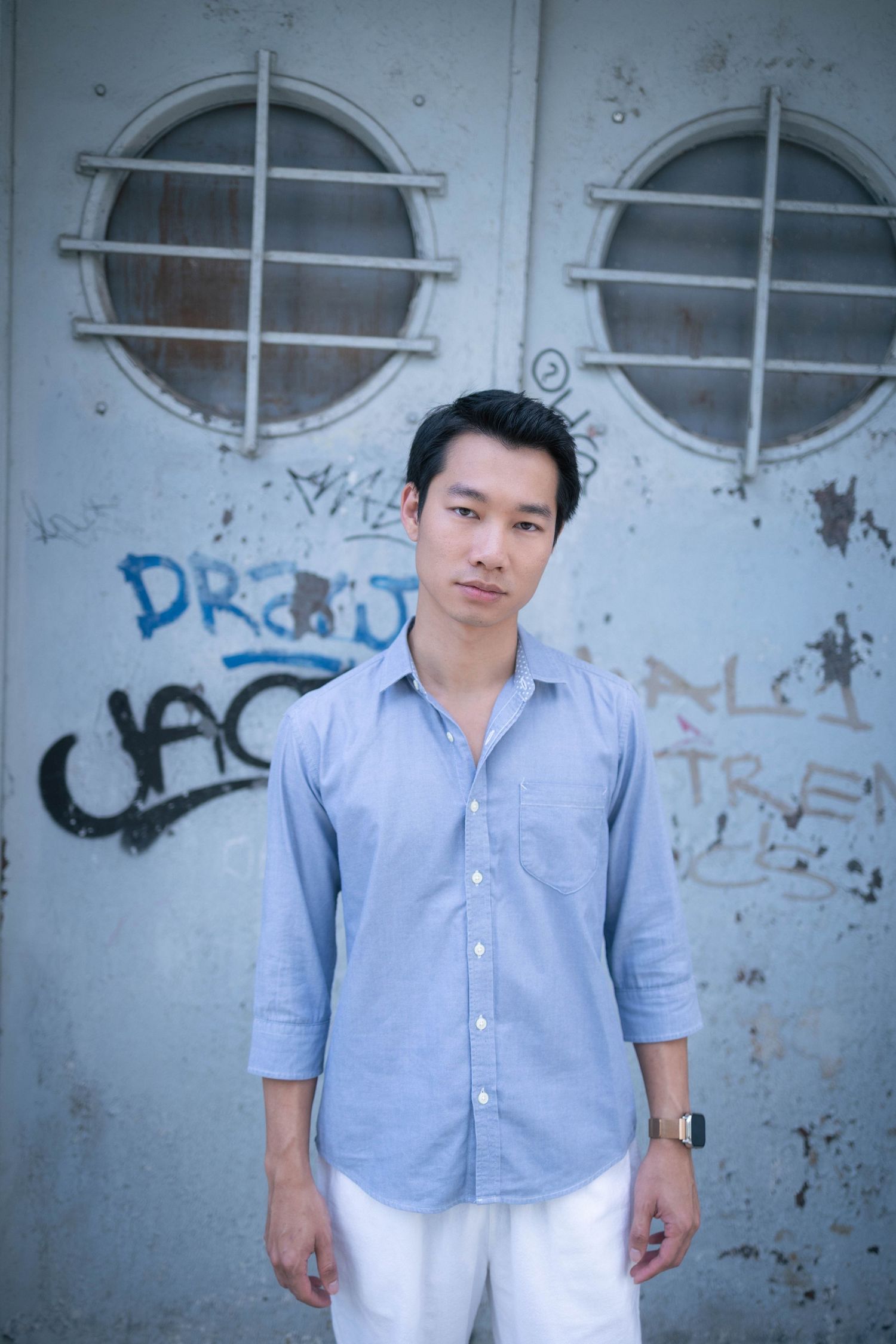 Young Asian man in blue shirt stands before weathered door with graffiti in urban setting.