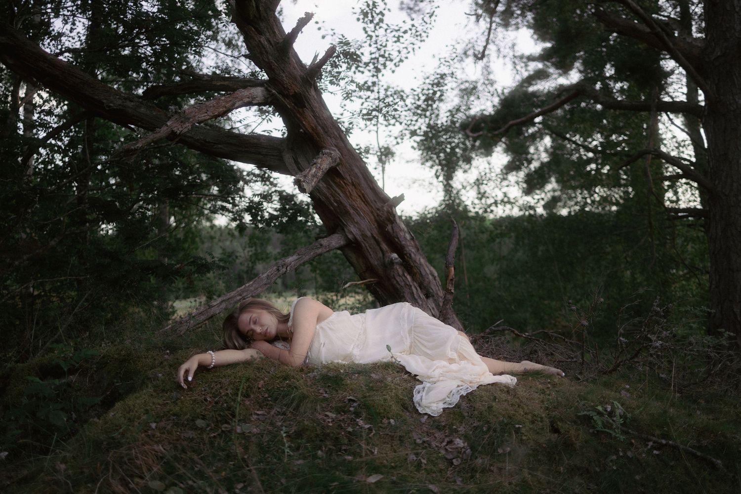 Woman in white dress lying peacefully beside fallen tree in forest setting.