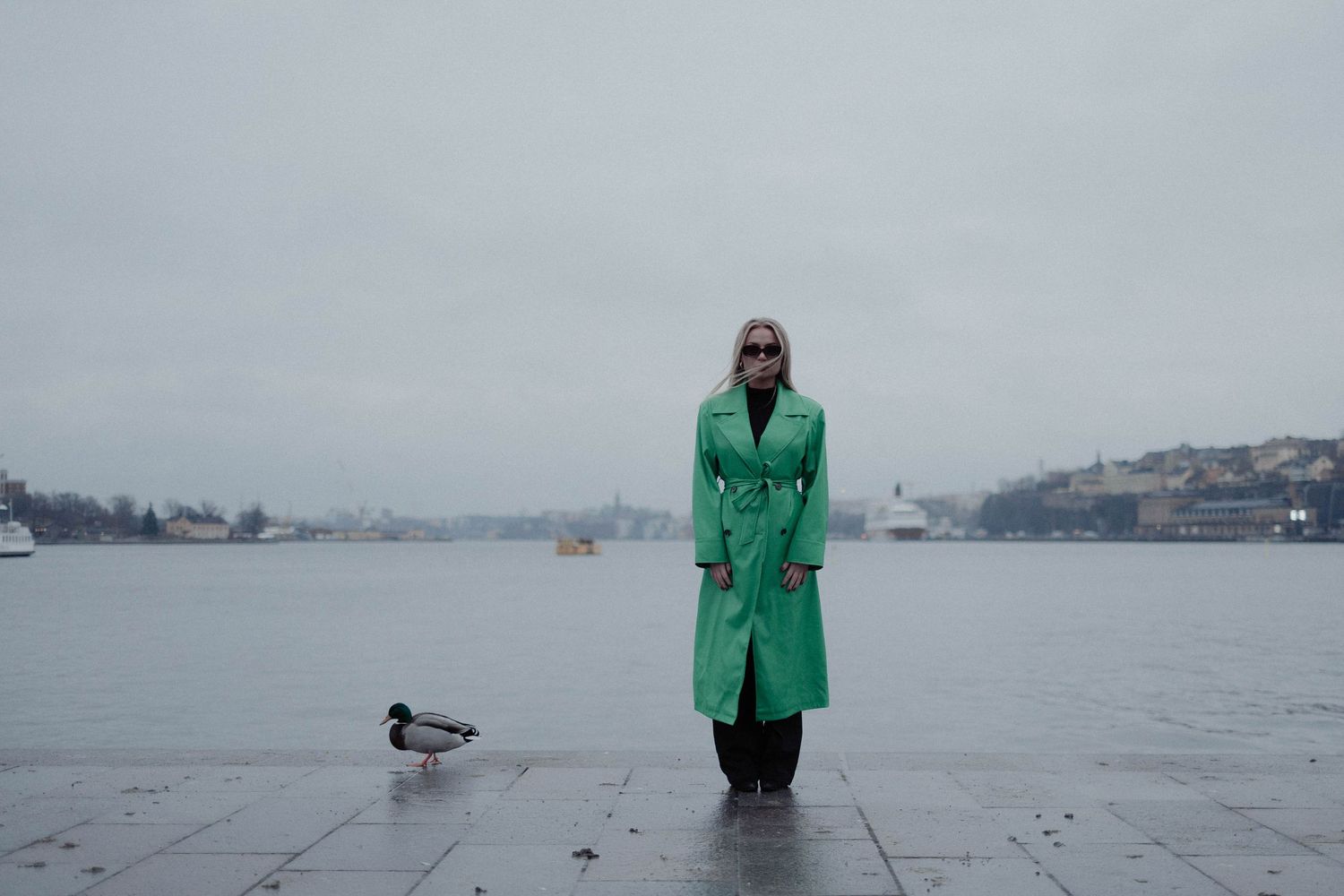 Woman in green trench coat standing by waterfront on overcast day with duck nearby.