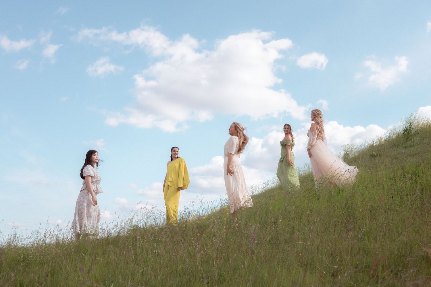 Five women in flowing pastel and yellow outfits stand on a green hillside under blue sky.