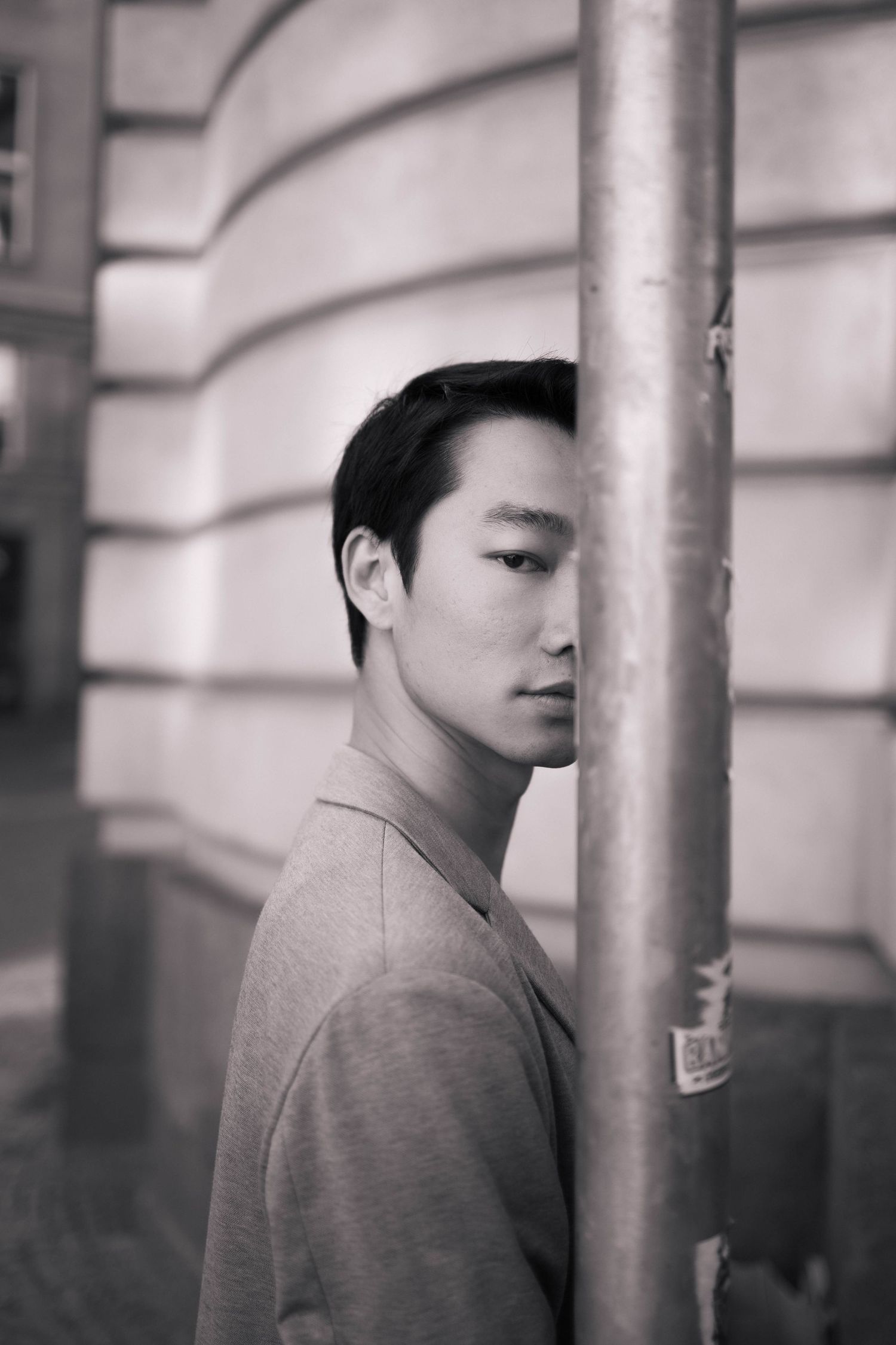 Black and white portrait of young Asian man leaning against metal pole on city street.