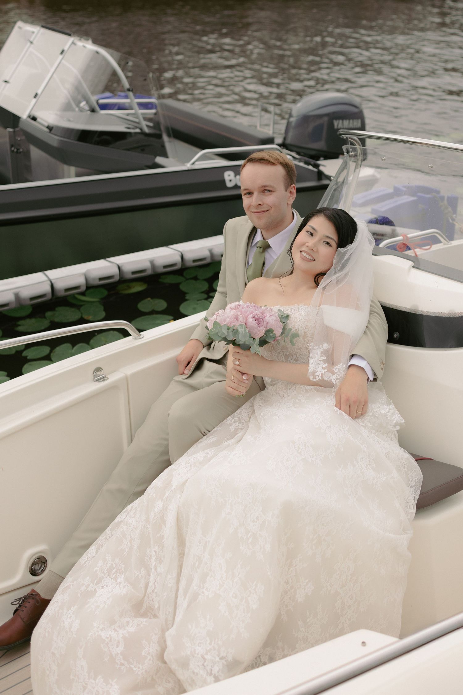 Bride and groom sitting together on a white boat, bride holding pink bouquet in lace wedding dress with veil.