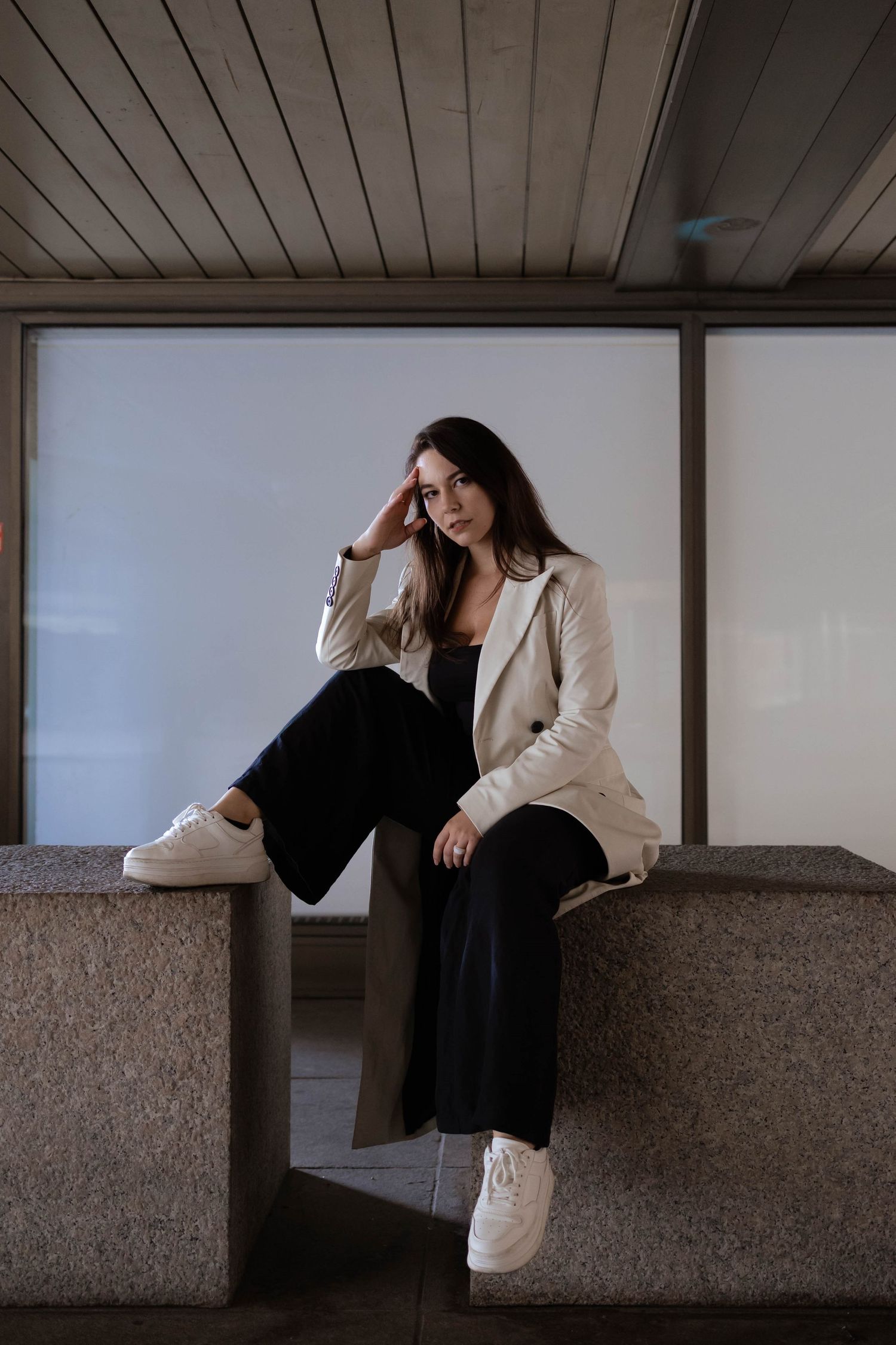 Young woman in beige blazer and black pants sits on concrete ledge in modern interior.