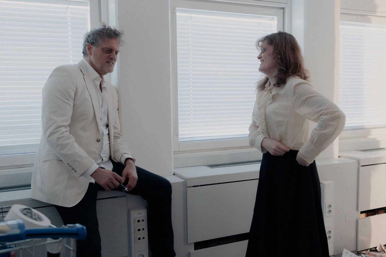 Man in white blazer and woman in white blouse having a conversation in a bright office setting.