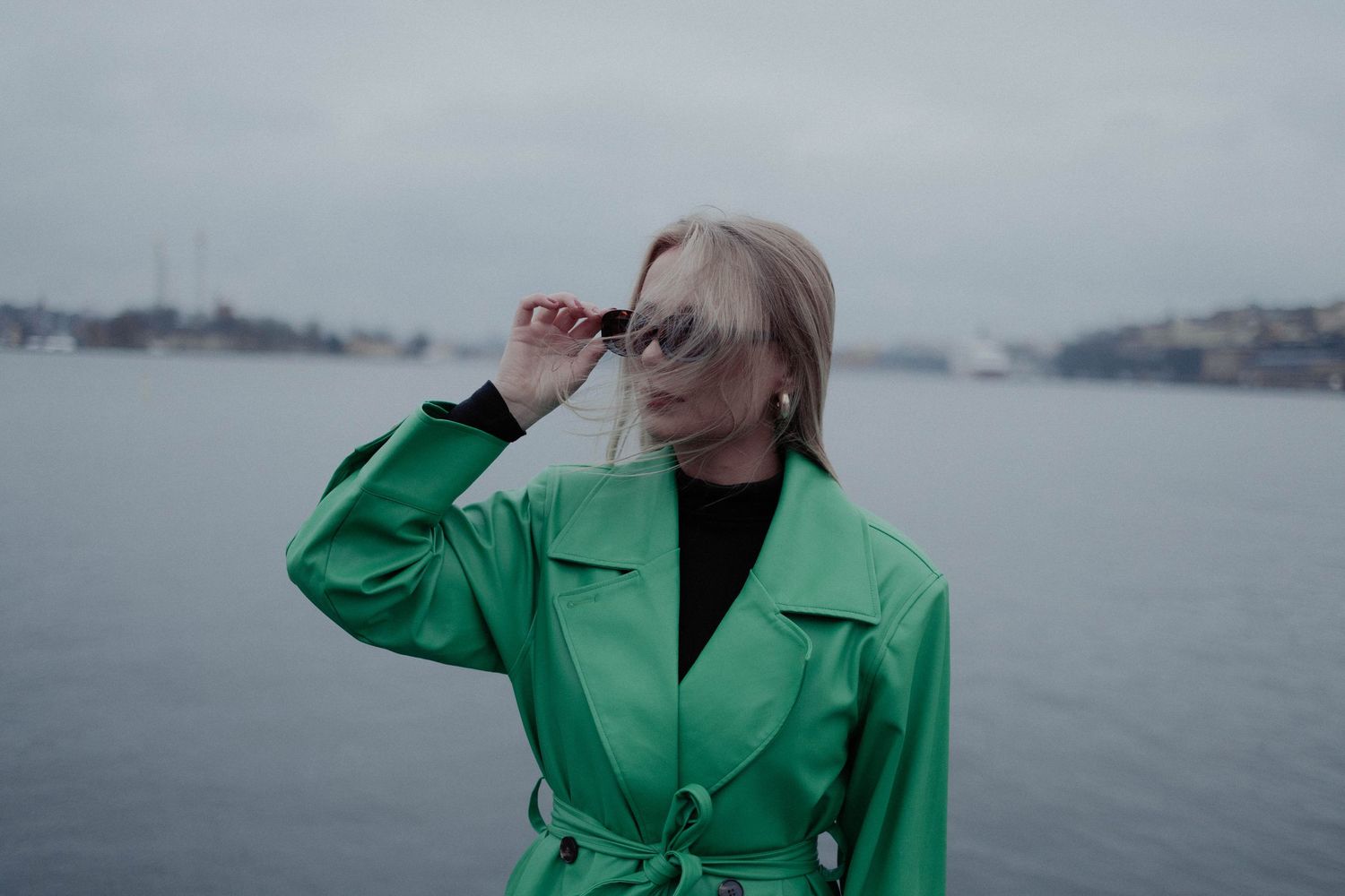 Blonde woman in a green leather coat poses by the water on a cloudy day.