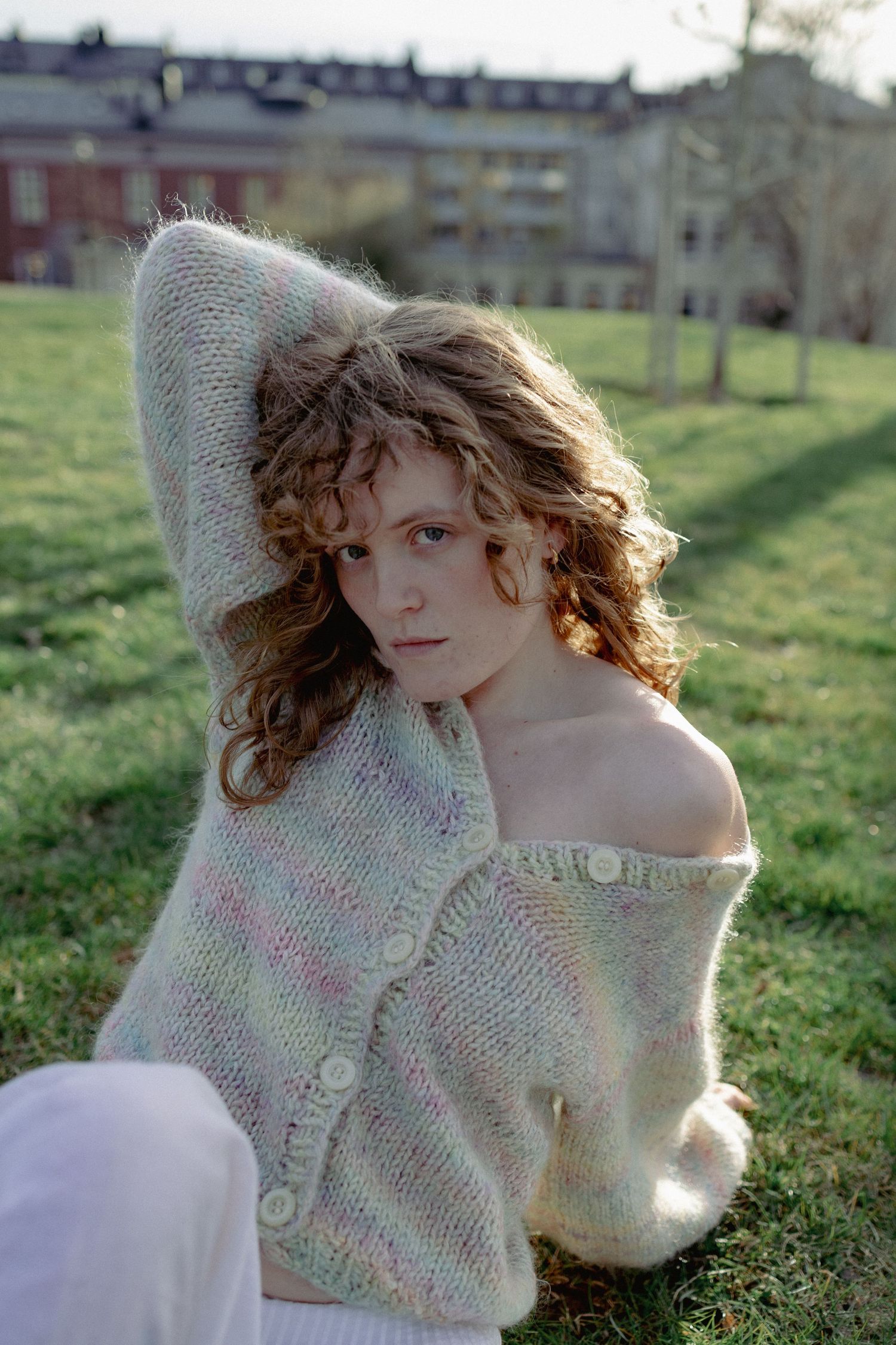 A curly-haired woman sits on grass in a pastel knit cardigan with one shoulder exposed in a park.