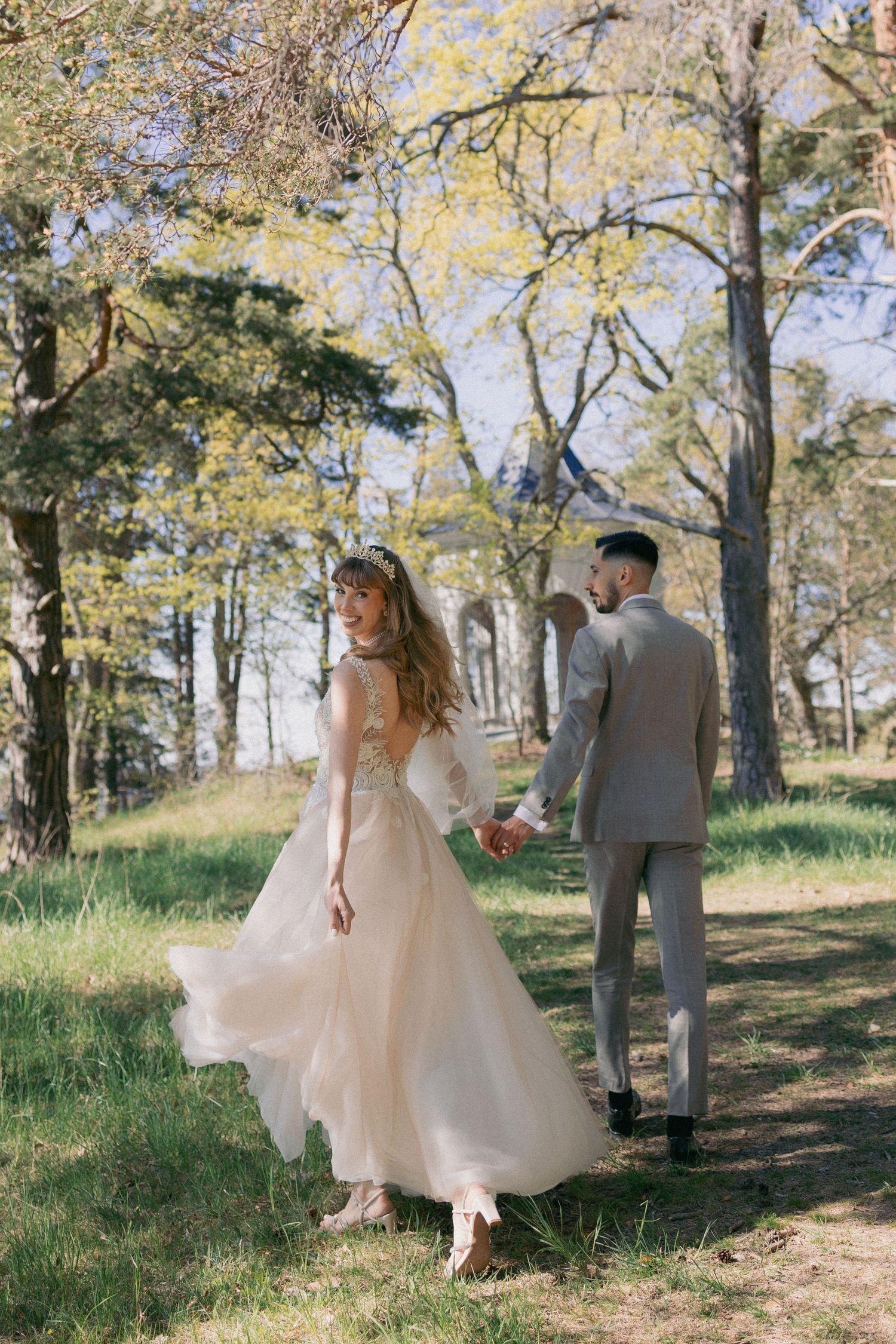 Bride in blush tulle gown smiling back at groom while walking through sunny woodland.