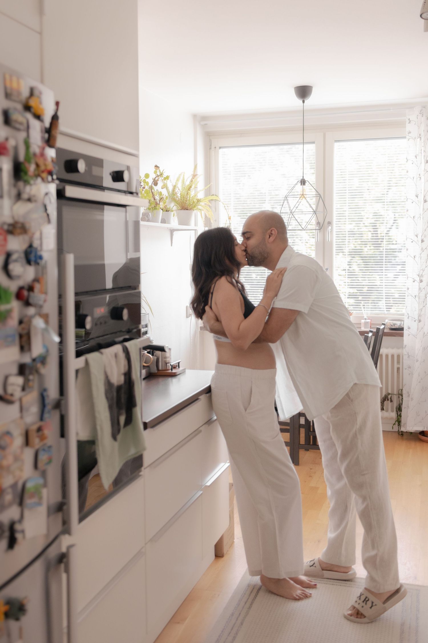 Couple sharing a kiss in a bright modern white kitchen, woman in black top and white pants.