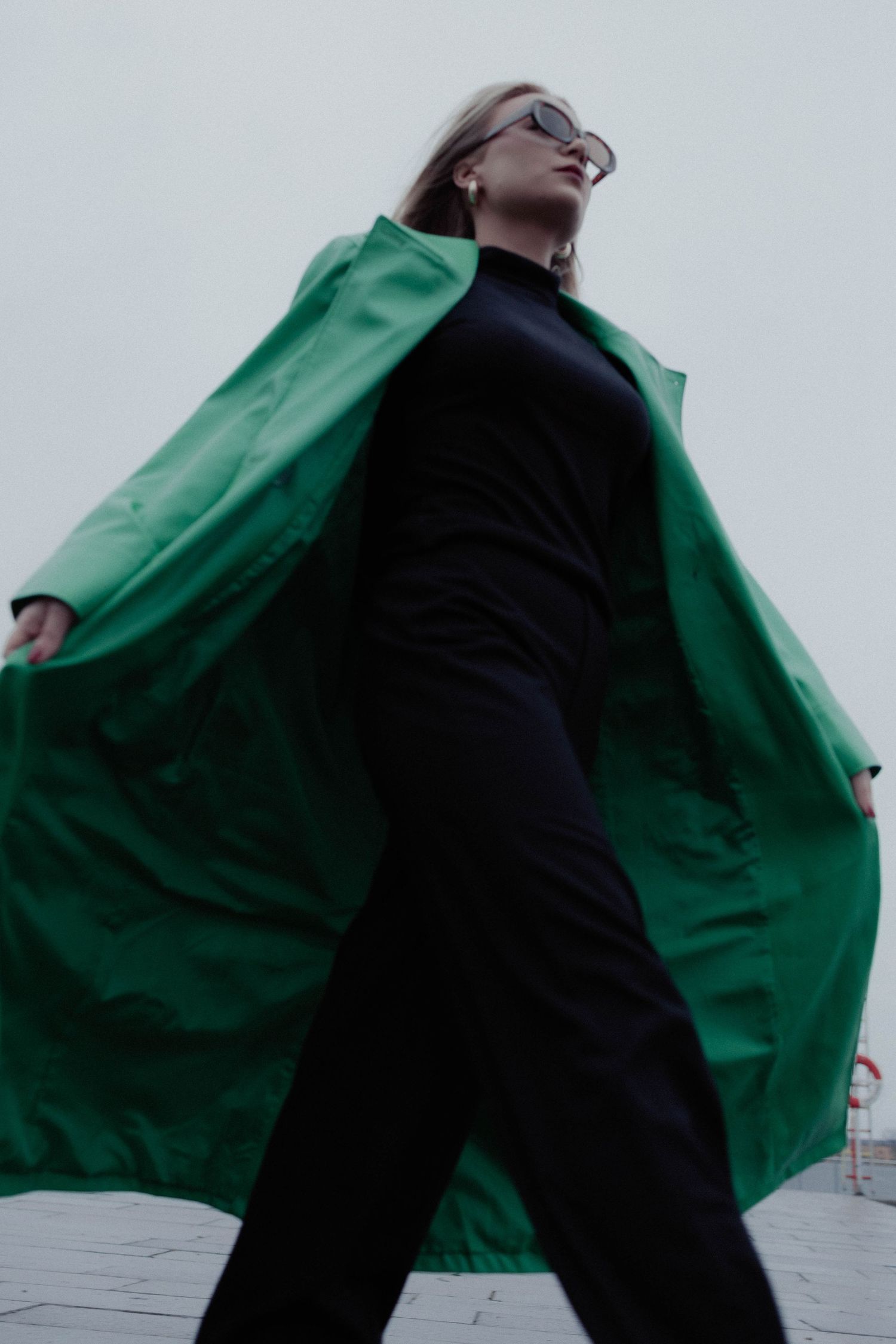 Low angle view of a blonde woman in a green coat and sunglasses posing confidently outdoors.