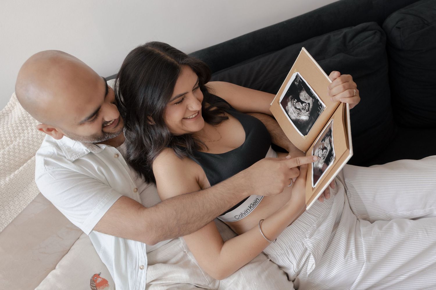 Couple lying on bed smiling at framed ultrasound photos, sharing a tender pregnancy announcement moment.