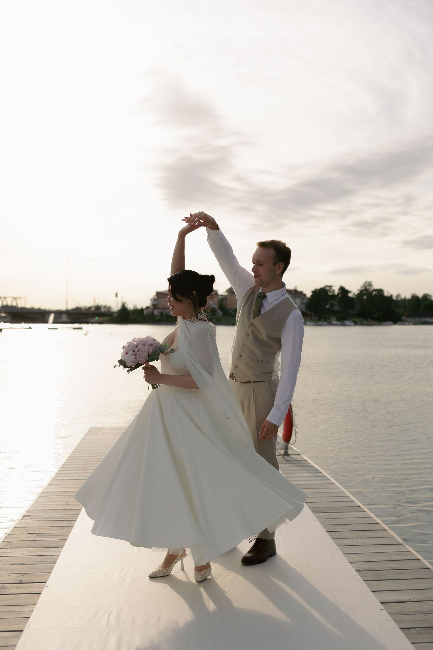 Bride and groom dancing on a white pier at sunset, bride twirling in tea-length dress holding pink flowers.
