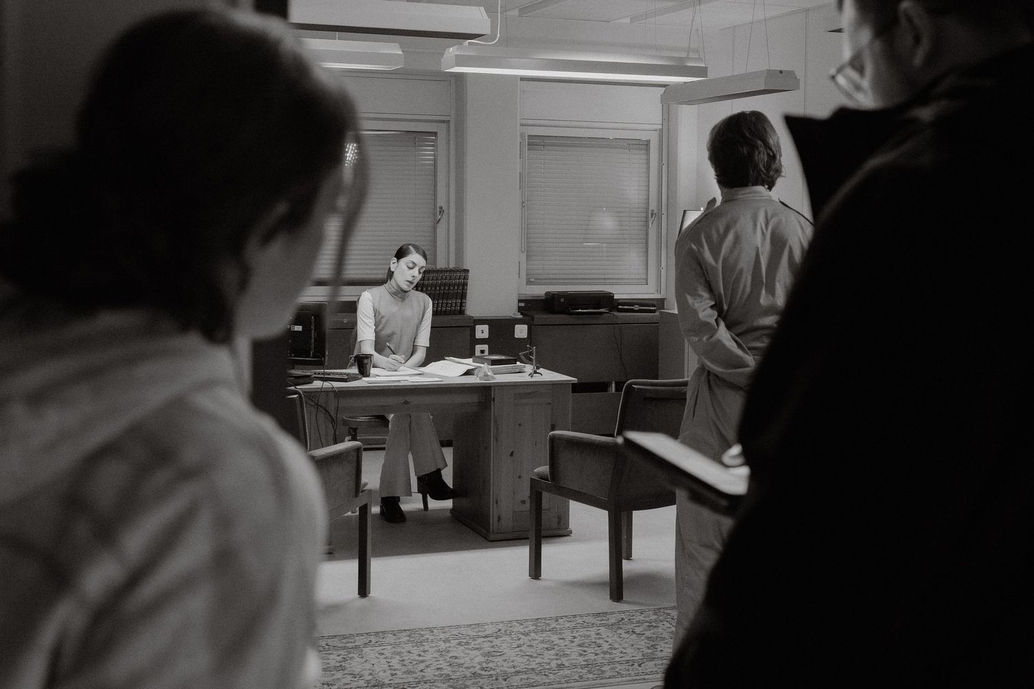 Black and white office scene with a woman at a desk and two people in the foreground.