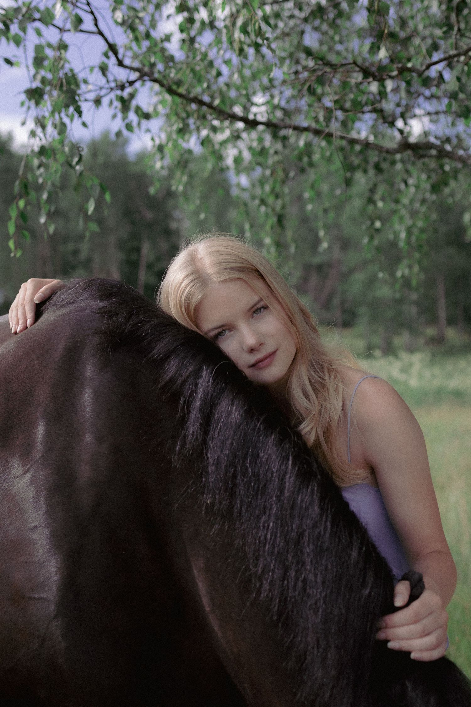 Blonde woman resting her head against a dark horse's mane in a green forest setting.