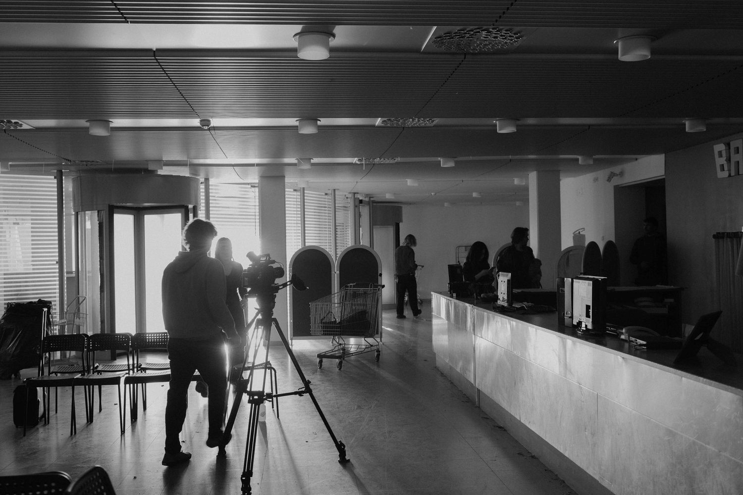 Cameraman filming in a dark office lobby with people gathered at a reception desk.