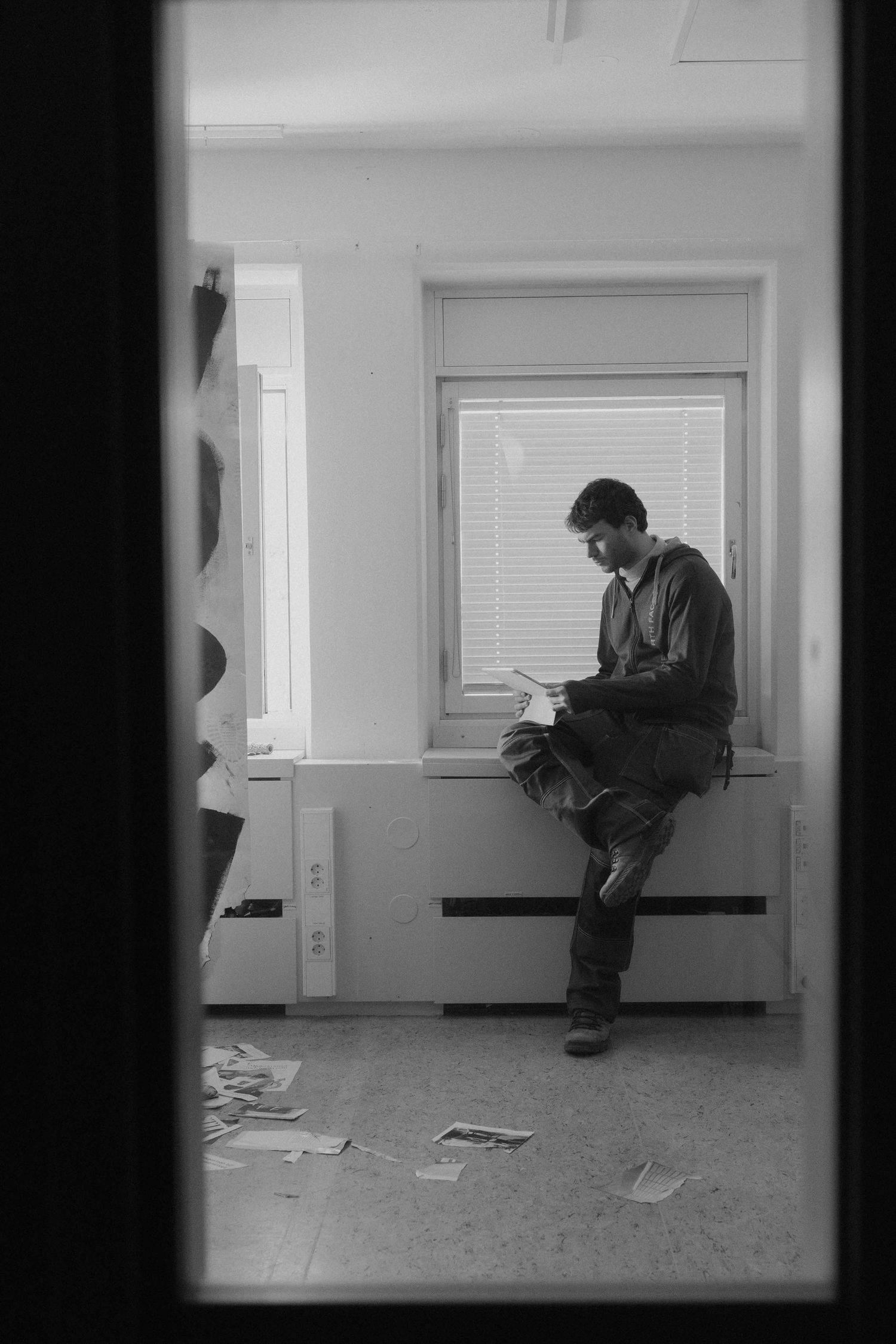 Black and white photo of man reading documents while seated on windowsill with papers on floor.