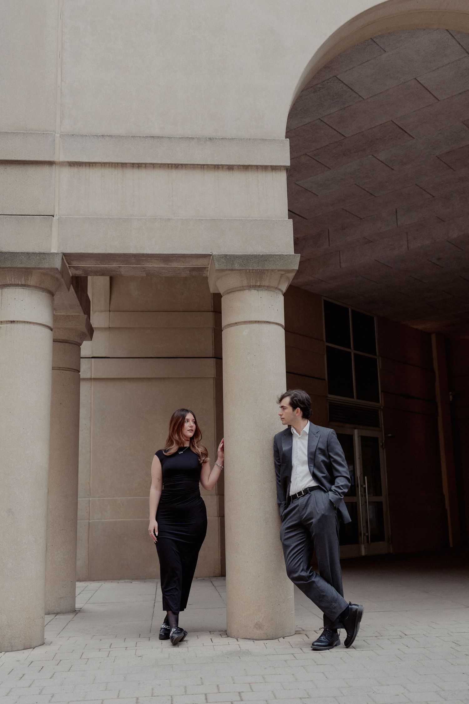 Couple in elegant black outfits standing between stone columns of a classical building, smiling at each other.