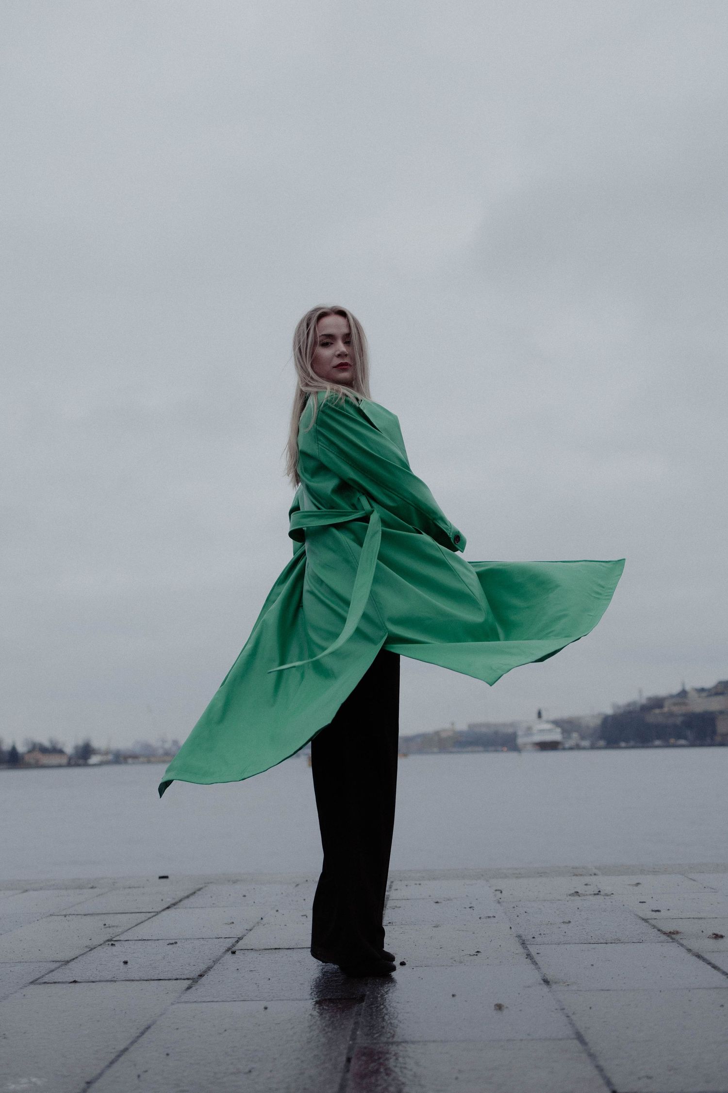 A blonde woman in a flowing green coat stands by a waterfront on an overcast day.