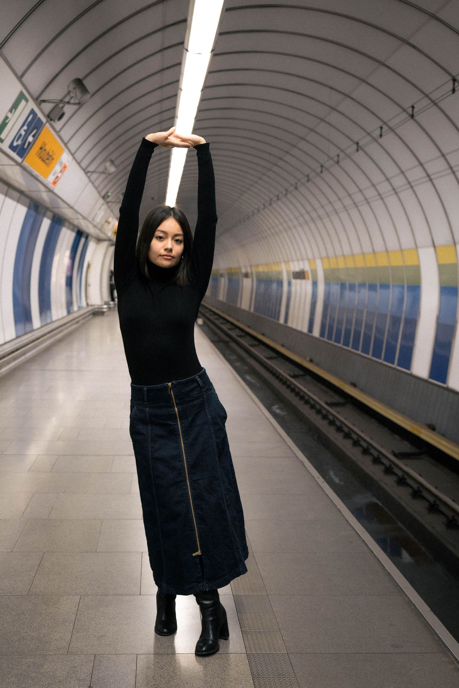 A young woman in black turtleneck and denim skirt poses with arms raised in a subway station tunnel.