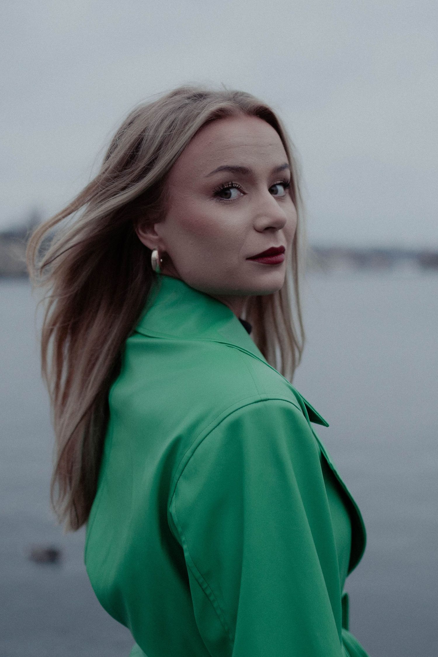 A blonde woman in a green leather jacket gazes back over her shoulder near a waterfront.