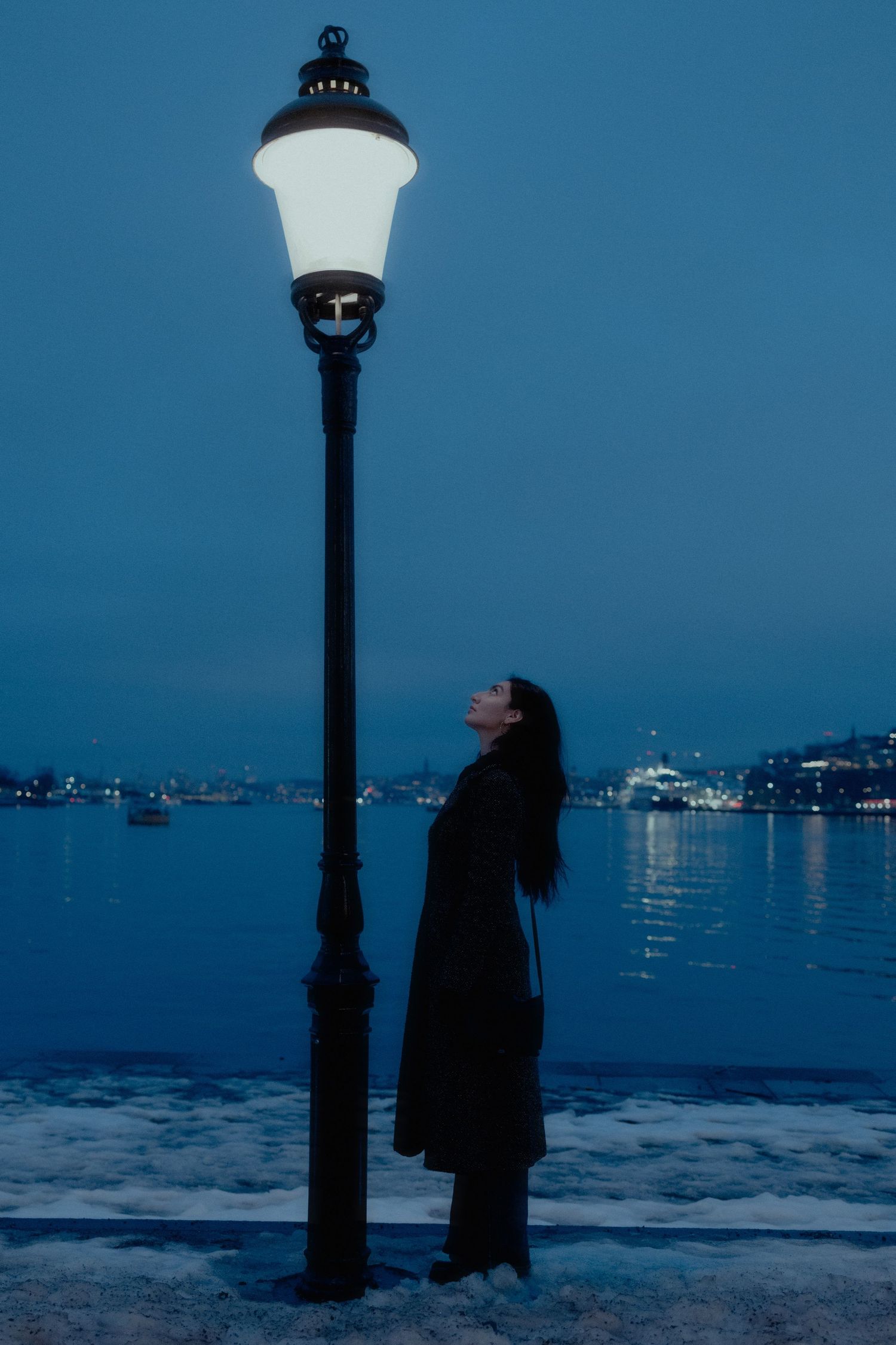 Woman in dark coat gazing up at glowing street lamp by frozen waterfront.
