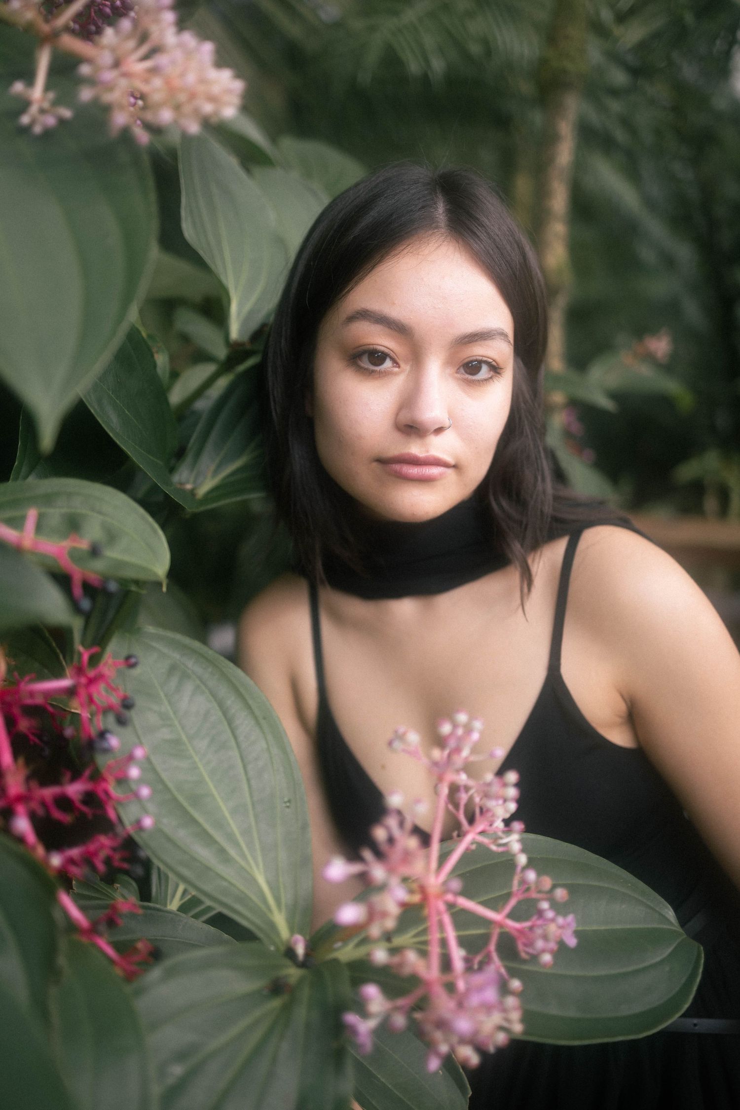 Young woman with black hair poses among tropical pink flowers and large green leaves.