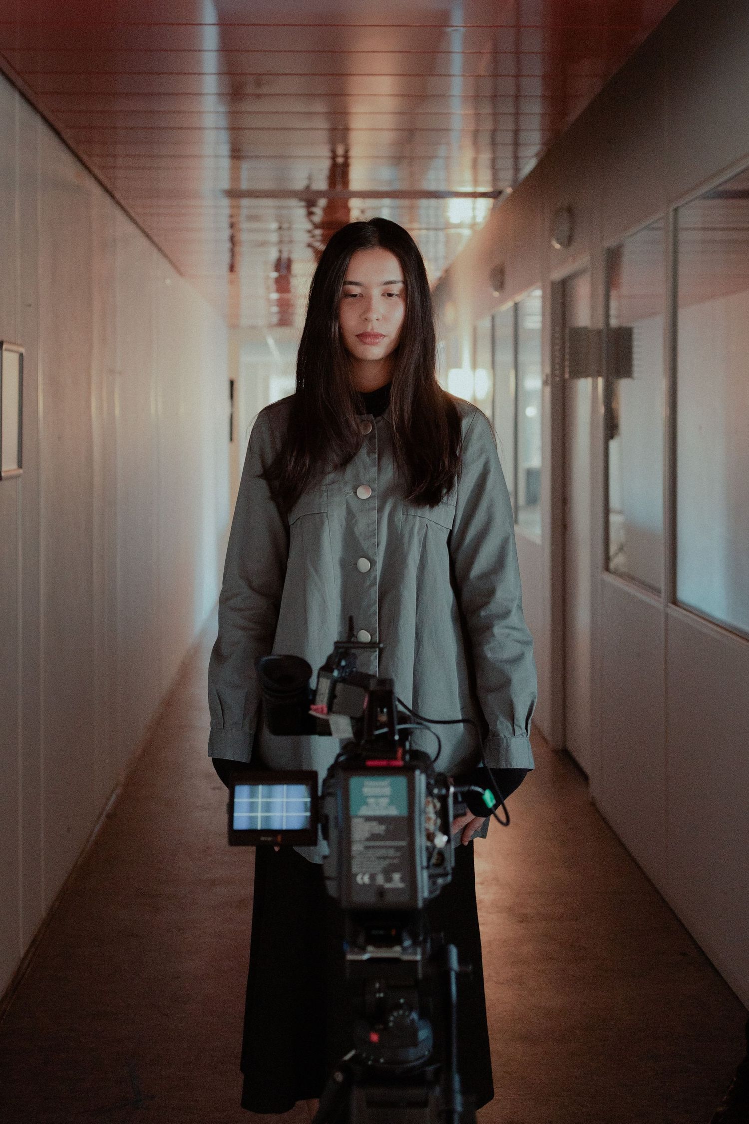 Young woman holding a professional cinema camera in a narrow dimly lit hallway.