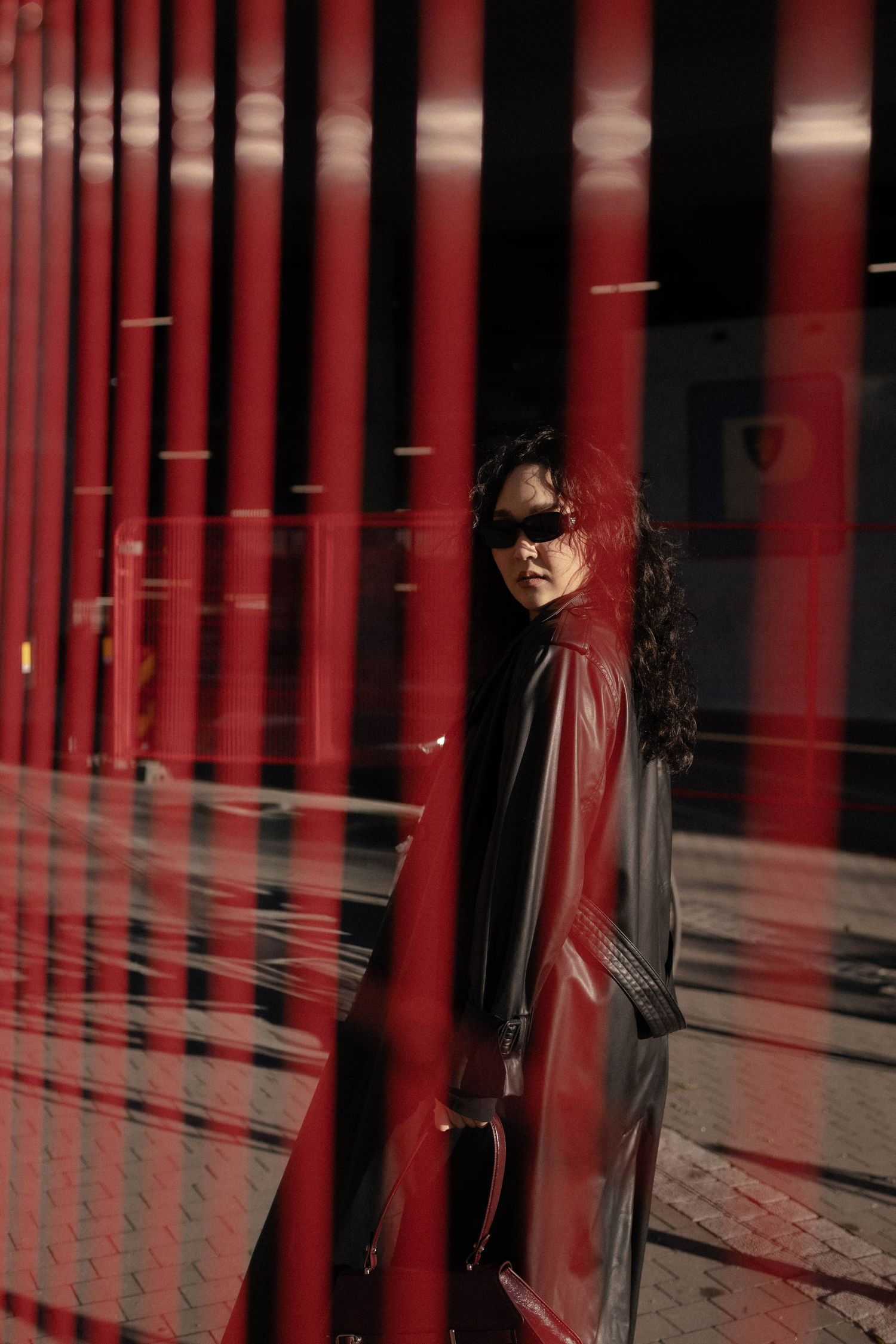 Woman in red leather coat and sunglasses poses against red pillars in dramatic urban setting.