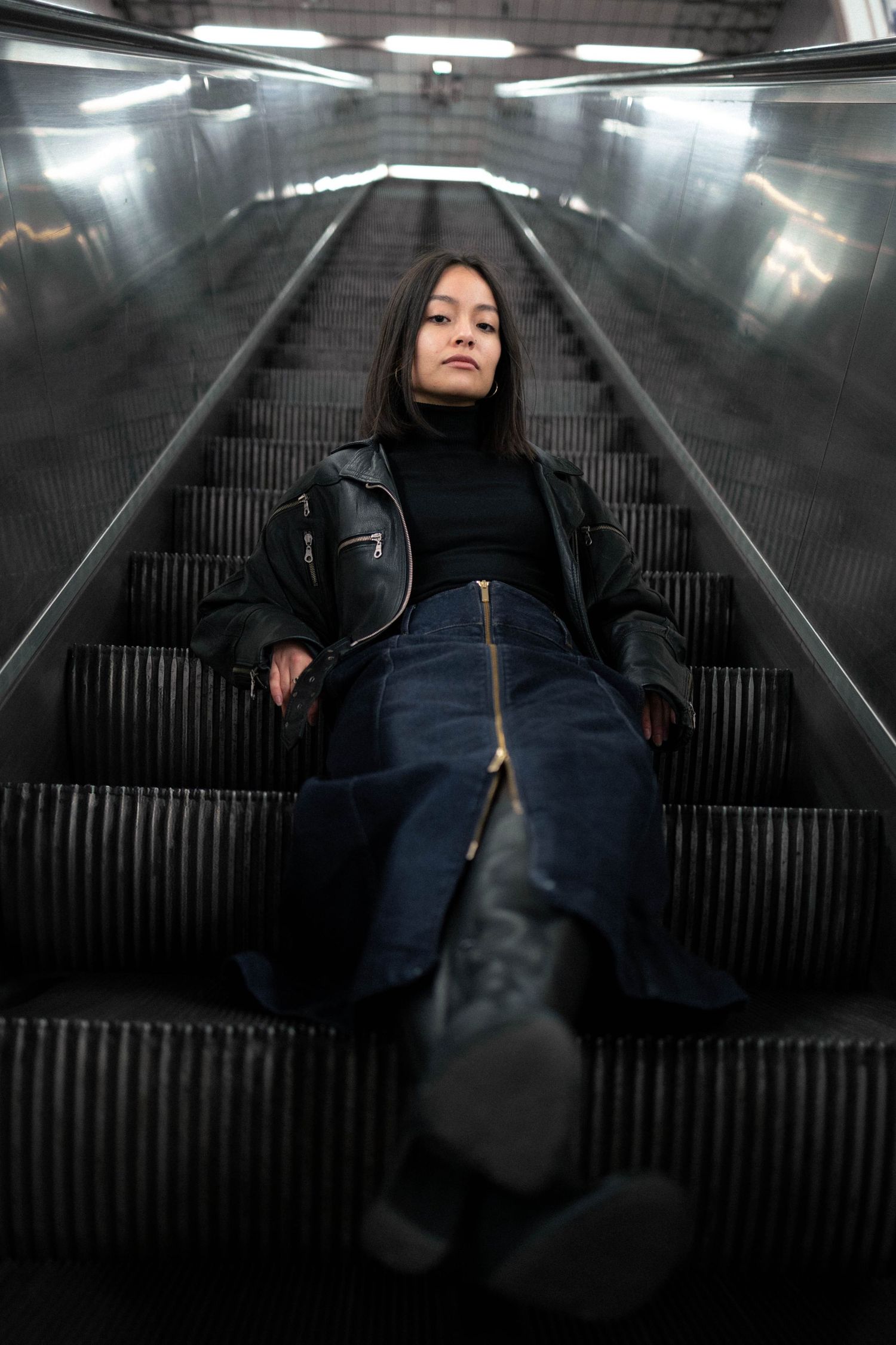 Young woman in leather jacket sitting on escalator looking at camera.