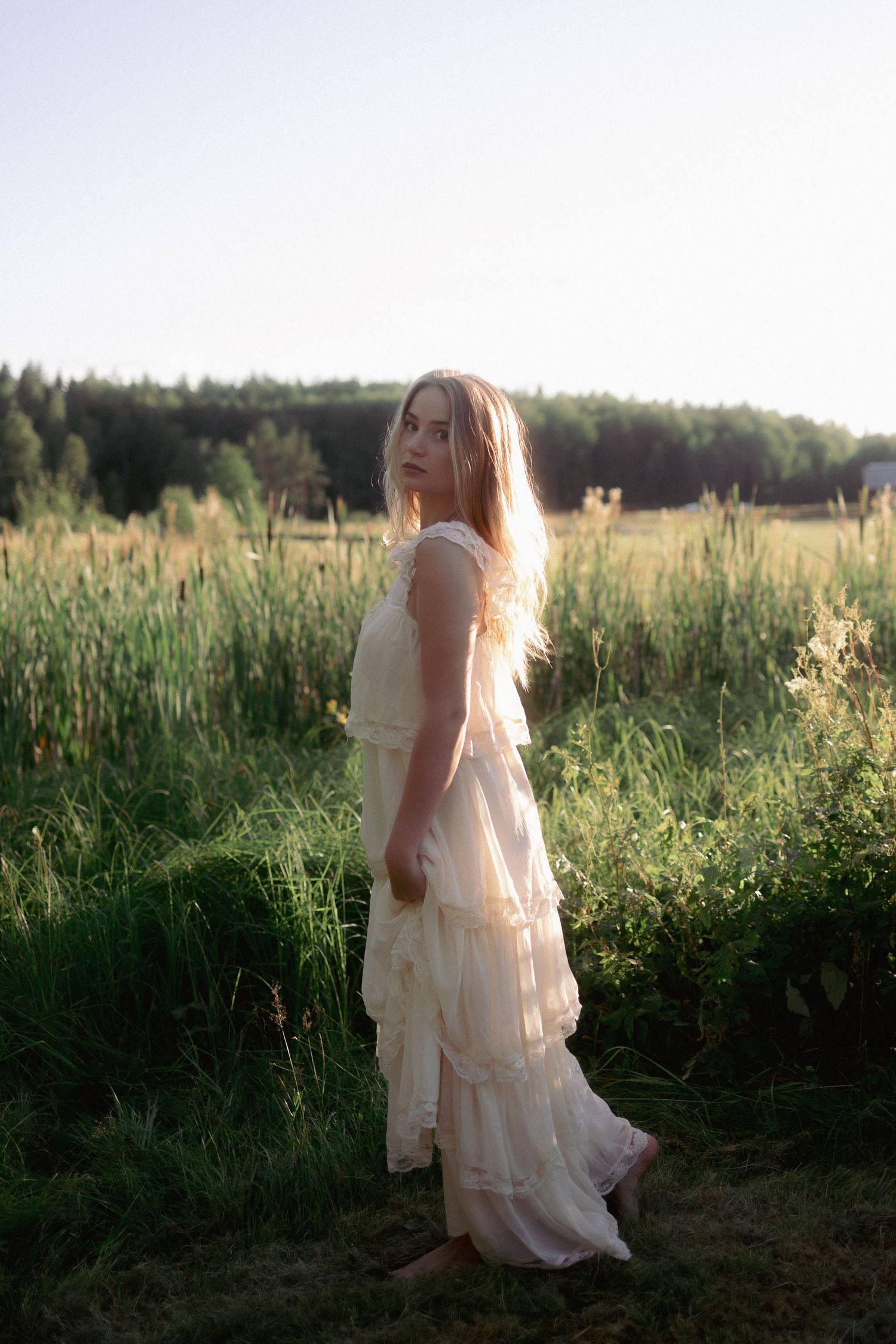 Blonde woman in flowing white tiered dress standing in a sunlit green meadow at dusk.