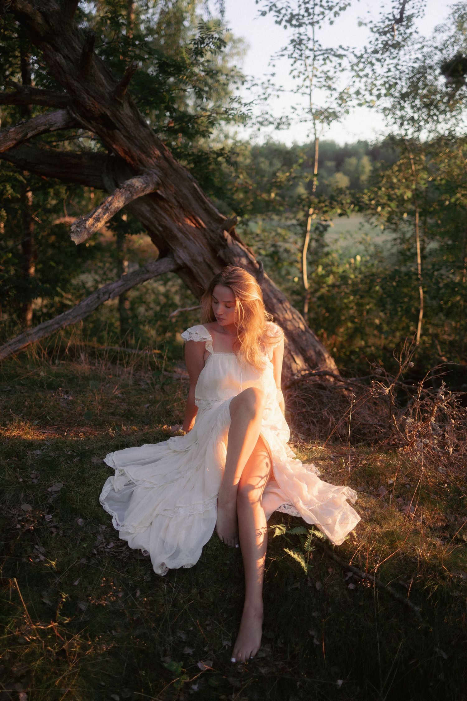 Blonde woman in flowing white dress sitting by fallen tree in warm sunset.