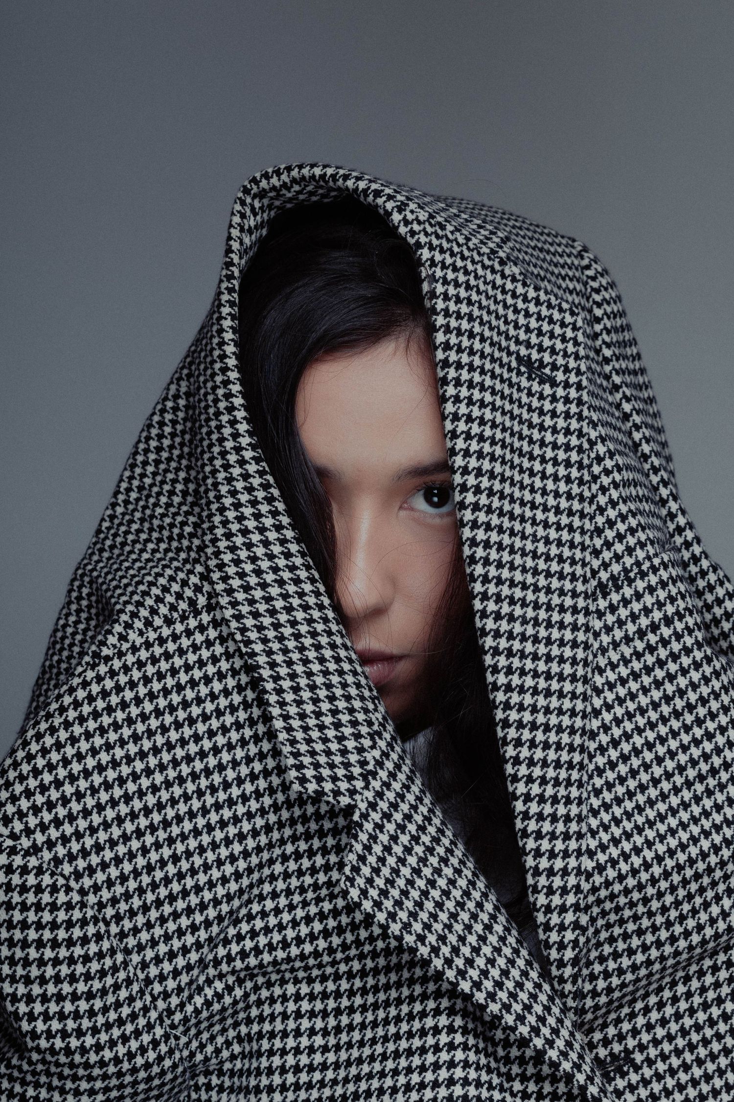 Person peeking through houndstooth patterned fabric against gray background.