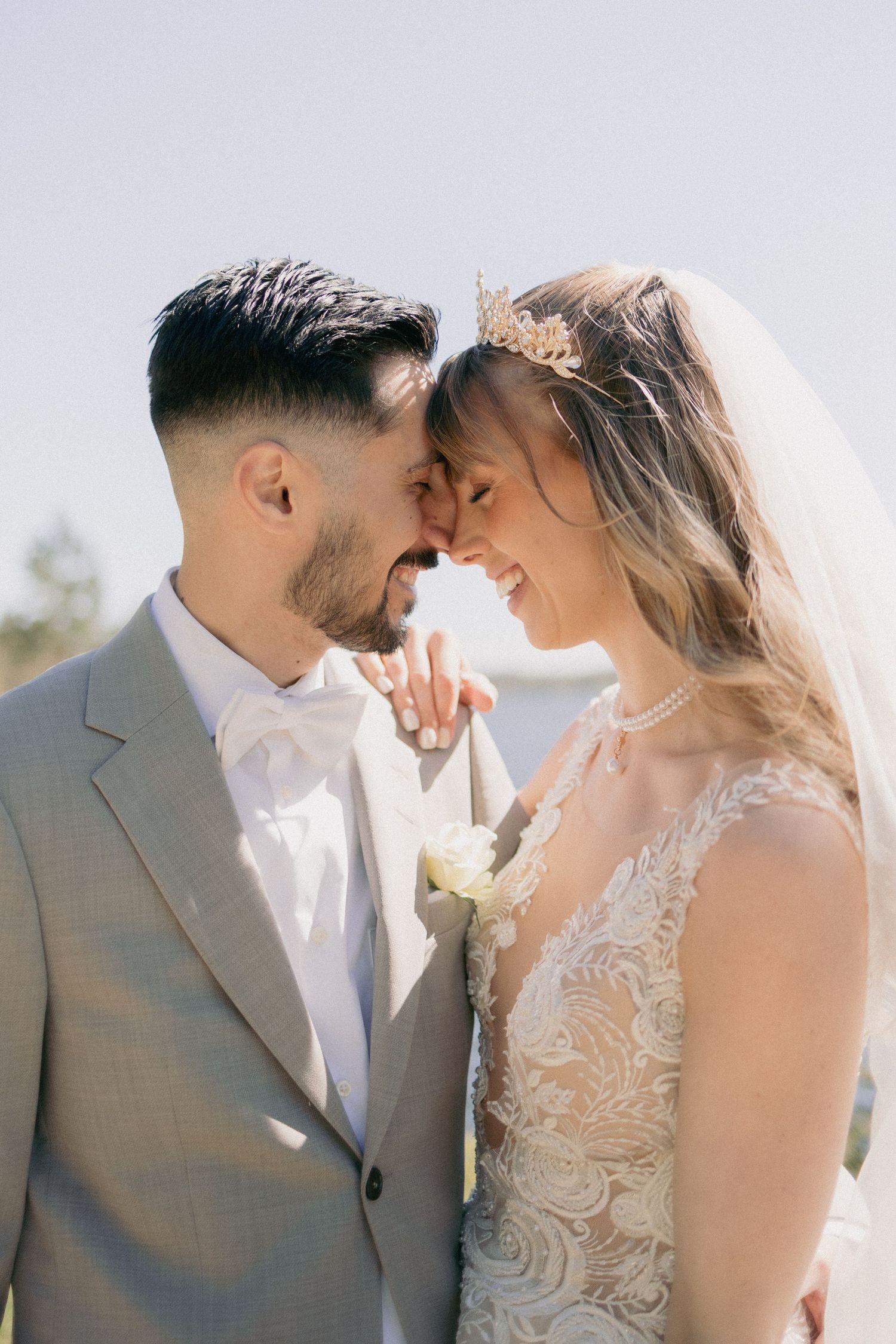 Bride and groom touching foreheads romantically outdoors, bride wearing lace dress with crown and veil.